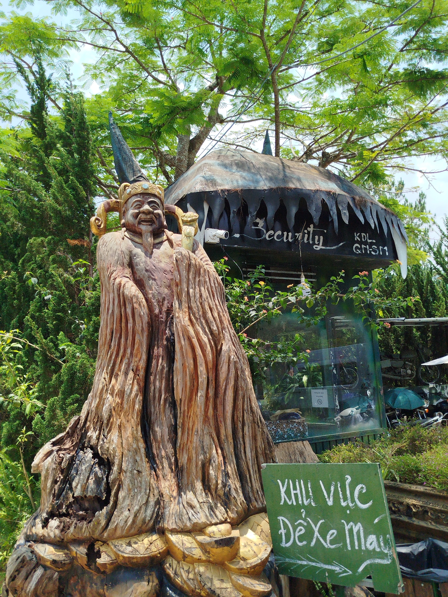 The image displays an outdoor scene dominated by a large, intricately carved wooden statue and a distinctive security booth, set amidst lush greenery under a clear, bright sky.
In the foreground, a tall statue of a smiling, jovial figure is carved from a tree trunk, featuring a beard, a detailed golden crown with light blue accents, and flowing robes that blend with the natural wood grain. The figure holds an object, possibly a staff, in its right hand.
Behind and to the right of the statue stands a unique security booth or hut. It has a dark, possibly metallic, roof with a jagged, tooth-like design along its edge. The word "Security" is painted in white script on its side, along with the letters "KDL GHSM". Below the roof, the booth features glass panels, revealing items like bicycles and an umbrella inside. A CCTV camera is visible near the roofline. Inside the glass, a "No Smoking" sign is present.
The entire area is surrounded by dense, vibrant green foliage, including tall coniferous trees and other deciduous trees with full canopies. The bright sunshine suggests it is midday under clear weather.
In the lower right corner, a green wooden sign, crudely painted in white, reads "KHU VỰC ĐỂ XE MÁY" (MOTORCYCLE PARKING AREA), with an arrow pointing to the right. This, along with the security booth, indicates a designated area, likely at a public attraction in Păng Tiêng 1, Vietnam. No specific activity is taking place other than the general presence of these installations.