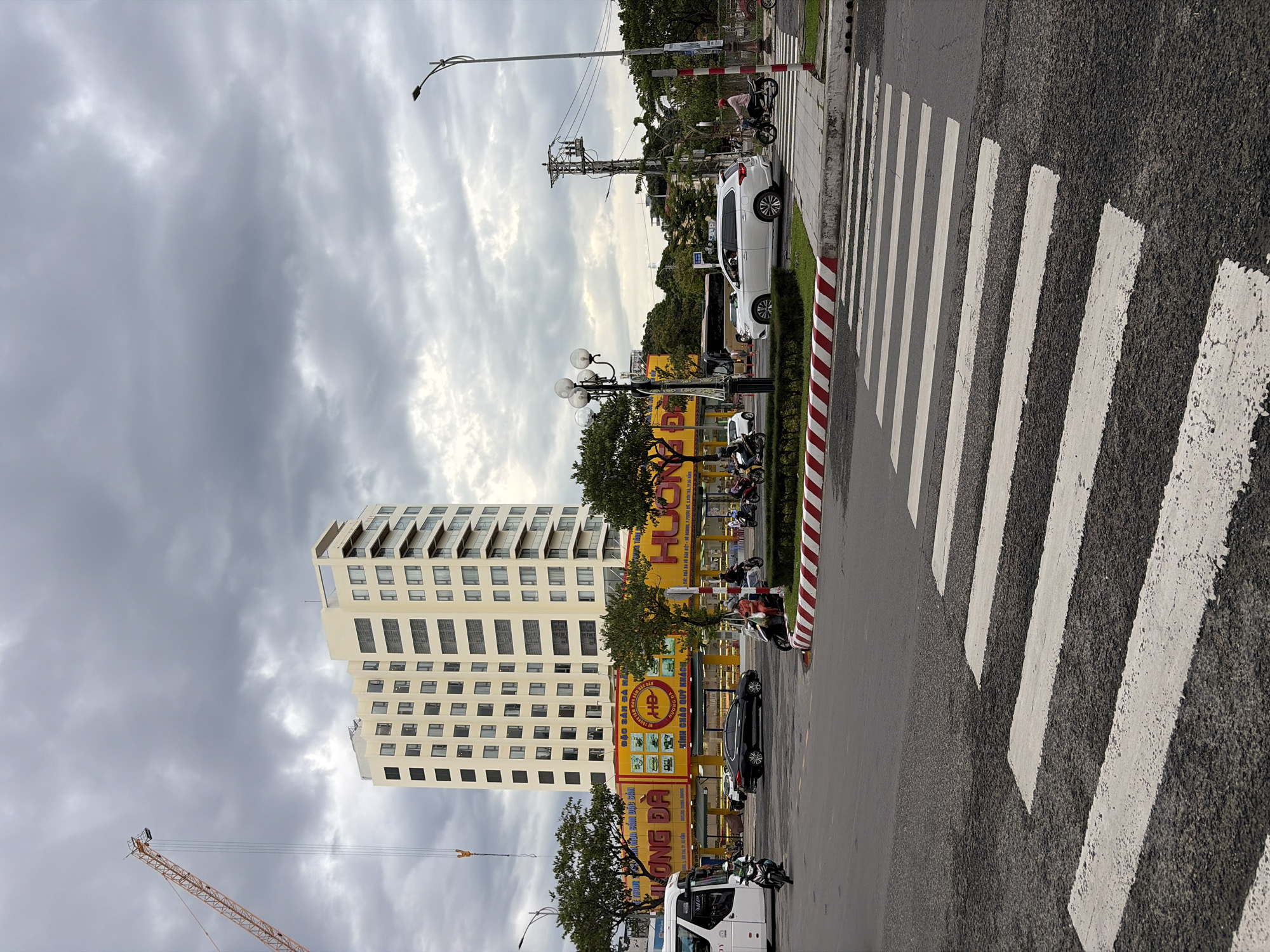 The image depicts a street scene in Đà Nẵng, Vietnam, under a cloudy sky. In the background, a tall, modern, cream-colored building with numerous windows stands prominently. To the left, a construction crane with an orange boom extends upwards against the overcast sky. At the base of the building, a bright yellow facade with red lettering, advertising "HUONG DA", is visible, suggesting a commercial establishment.
The foreground shows a road with a marked crosswalk. Several vehicles are present on the street, including a white bus on the left, a black sedan, and a white SUV. Motorcyclists are also visible, with one riding a white motorcycle in the foreground, another on a dark motorcycle near the yellow building, and a few others further in the background. Pedestrians are present on the sidewalk near the crosswalk and the yellow building. The street is lined with trees and a lamppost. The scene suggests an active urban environment, likely a commercial district within the city.
