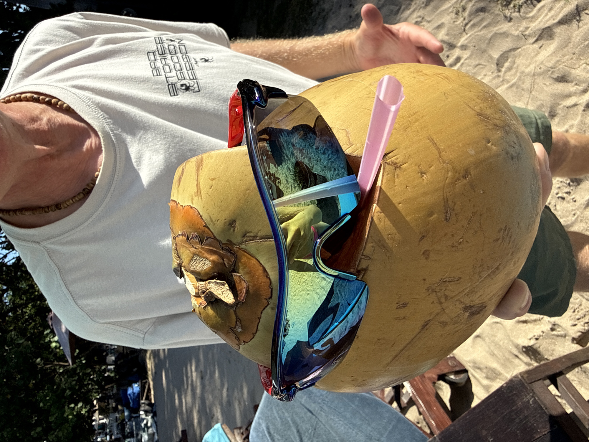 A person holds a large, golden-brown coconut with a pink straw inserted into it. The coconut is adorned with a pair of blue and red sunglasses resting on top, reflecting the surroundings. The person is wearing a white t-shirt with a graphic logo on the front, a wooden beaded necklace, and dark green shorts. The scene is set on a sandy beach, likely in Denpasar, Indonesia, with hints of trees and beach chairs in the background. The lighting suggests a sunny day.