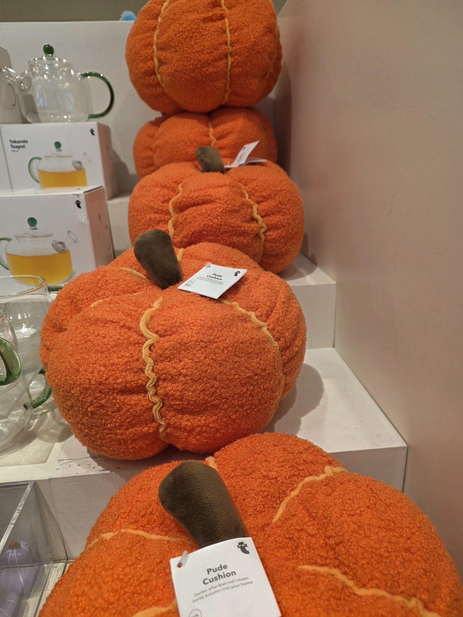 In a retail setting, likely a home goods store, a stack of plush pumpkin-shaped cushions is displayed on white shelves. The cushions are various sizes and are all a vibrant orange color with textured fabric and decorative yellow stitching resembling pumpkin ribs. One cushion at the bottom has a tag with the text "Pude Cushion" visible. To the left of the cushions, a glass teapot with a green handle and lid is partially visible, along with a glass cup. The background is a plain, light-colored wall. The scene is well-lit and focuses on the seasonal decor.