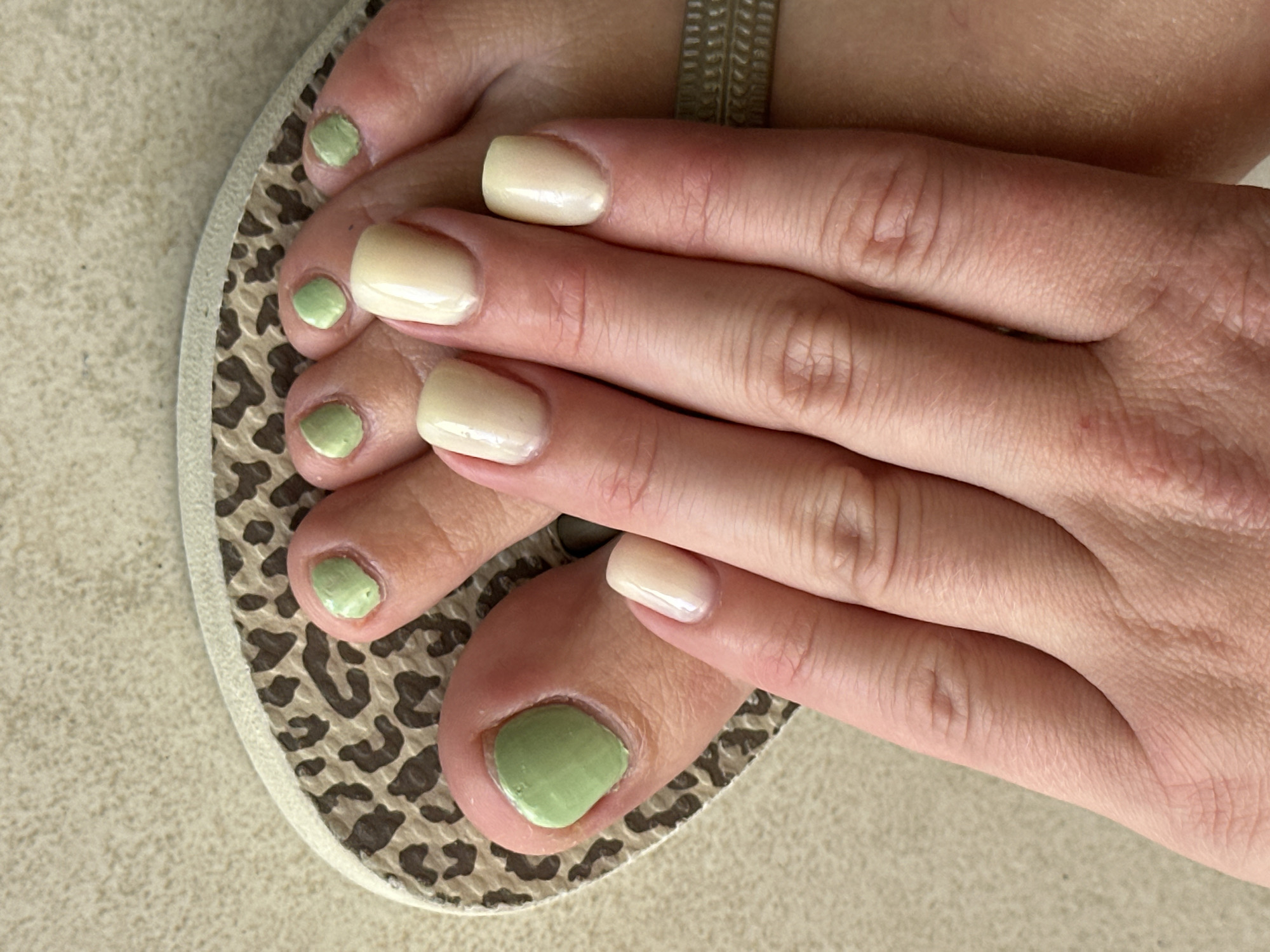 A top-down shot depicts a person's left foot in a flip-flop, with their hand resting on their toes. The fingernails are painted with a light, almost off-white color, while the toenails are painted in a light green shade. The flip-flop has a neutral-toned leopard print design. The background is a plain, light beige surface. There is no evidence to indicate this scene is located in Petah Tikva, Israel.