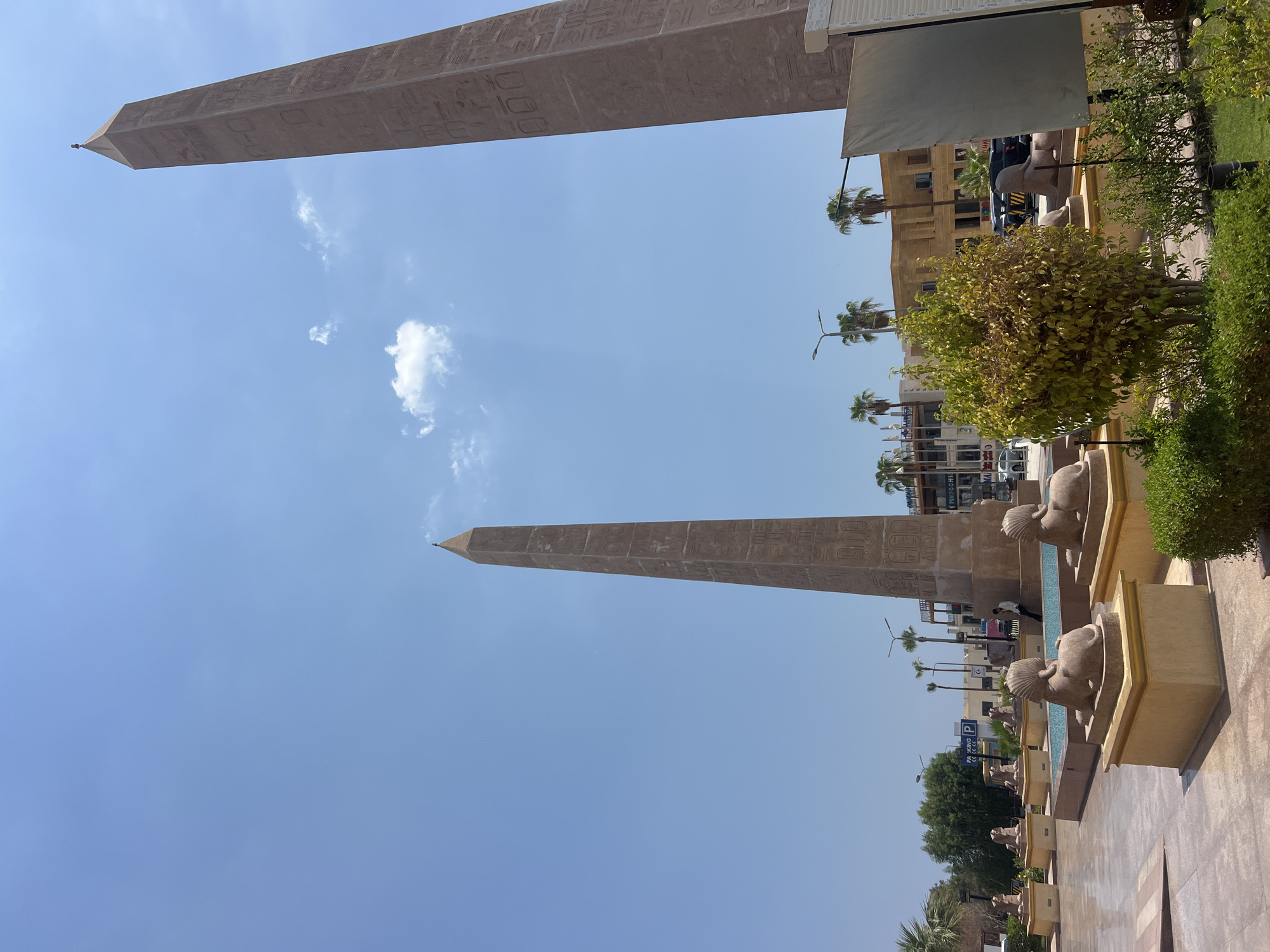 This is a daytime, eye-level, outdoor photograph of Naama Bay in Sharm El Sheikh, Egypt. The image captures two large, ancient Egyptian-style obelisks, one in the left foreground and another in the right midground. Both obelisks are inscribed with hieroglyphs and are surrounded by a plaza. In front of the obelisks are several sphinx statues, some of which are positioned next to a shallow pool of water. Palm trees are visible in the background, along with several buildings that appear to be shops or restaurants, and a clear blue sky with a few scattered clouds. The overall scene depicts a public space with a prominent display of Egyptian heritage.