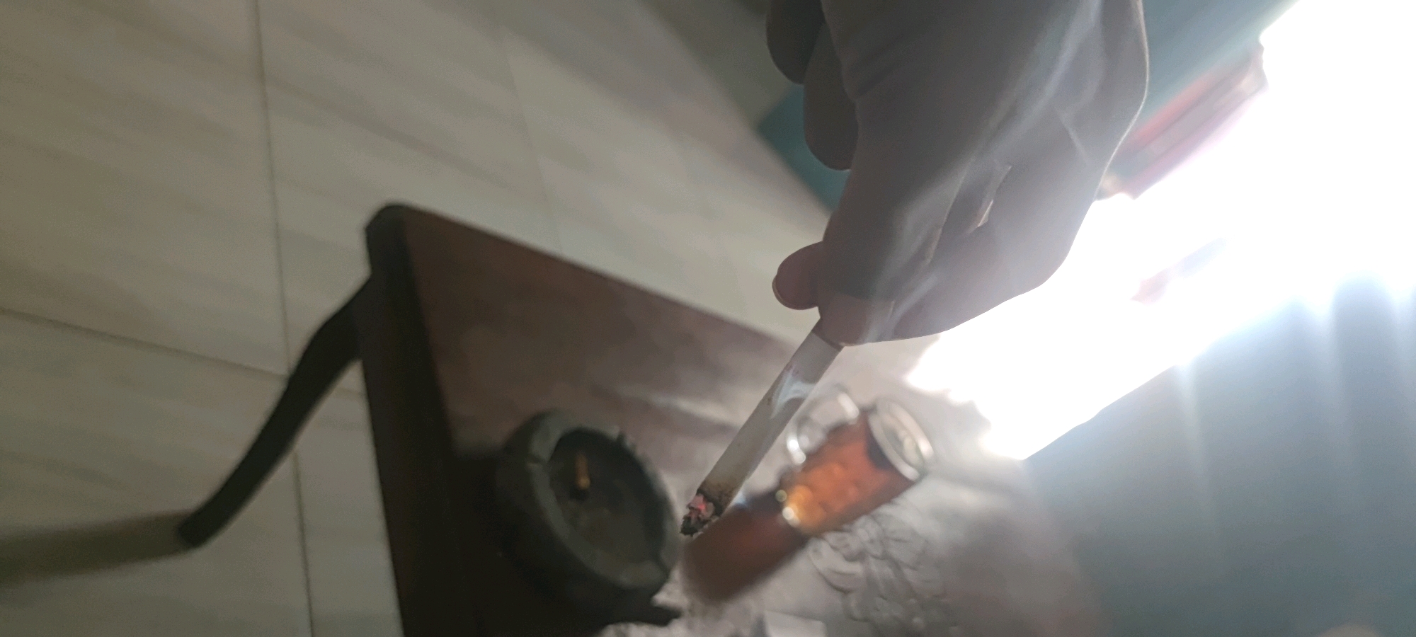 In Gambarsari, Indonesia, a close-up shot from an angled perspective shows a person's hand holding a lit cigarette, with smoke rising. The cigarette is positioned over a small, dark ashtray and a glass containing a dark liquid. The scene is illuminated by bright, overhead light, creating lens flares and washing out parts of the background. A portion of a wooden surface, possibly a table, is visible beneath the objects. The overall impression is one of a dimly lit interior with harsh lighting.