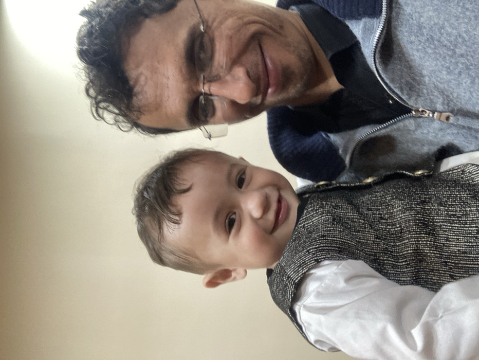 A man with glasses and curly hair is taking a selfie with a smiling toddler. The man is wearing a gray zip-up sweater over a dark blue shirt. The baby is wearing a white shirt with a textured, dark-colored vest. The camera is angled downwards, focusing on their faces. The background is a plain, light-colored wall.