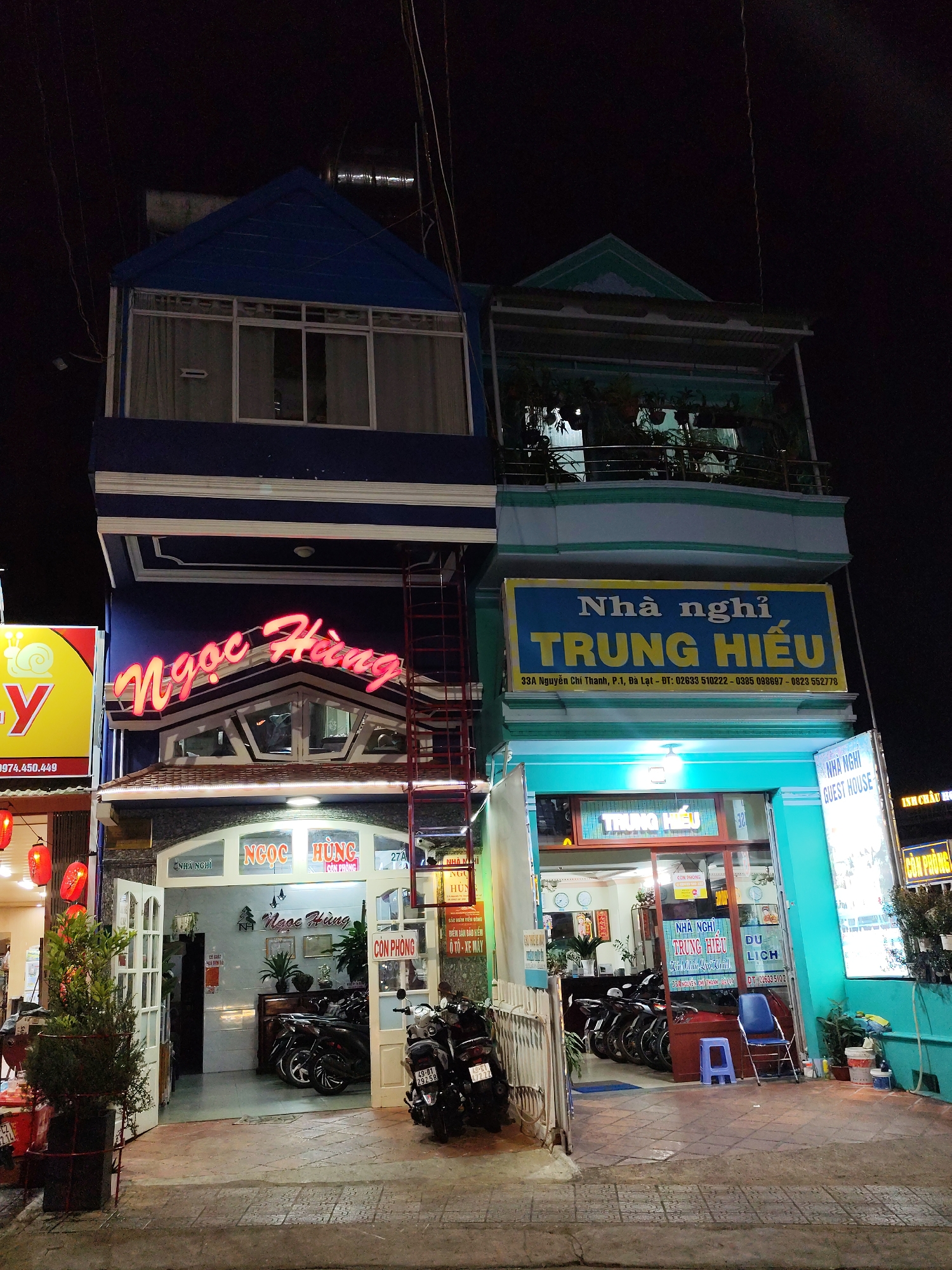 The image depicts the facades of two adjacent guesthouses, "Ngoc Hung" and "Trung Hieu," at night in Da Lat, Vietnam. "Ngoc Hung" has a blue and white exterior with a red neon sign. A ladder is leaning against the building. Motorbikes are parked in front of both guesthouses. "Trung Hieu" has a blue and teal exterior. The signs include phone numbers and addresses of each. Red lanterns adorn the side of one building.