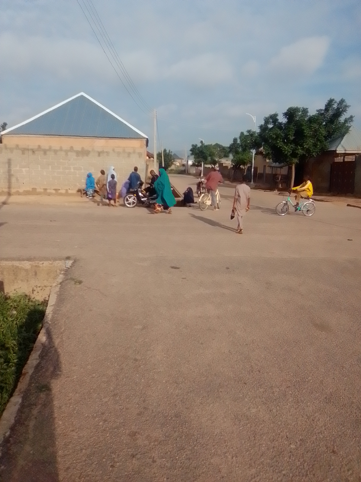 The image depicts a street scene in Liji Hill, Nigeria. A group of individuals are gathered around a motorcycle on the right side of the road. Some are standing, while others appear to be seated or leaning. One person is wearing a bright turquoise garment, and another is in a yellow shirt. A person on a bicycle, wearing a yellow shirt, is in motion further down the road to the right. On the left side of the road, a man in a light brown robe walks away from the viewer, carrying a light-colored object. Further left, several people are standing near a building constructed with light-colored bricks and topped with a blue corrugated metal roof. The road surface is asphalt. The environment includes scattered trees and a cloudy sky.