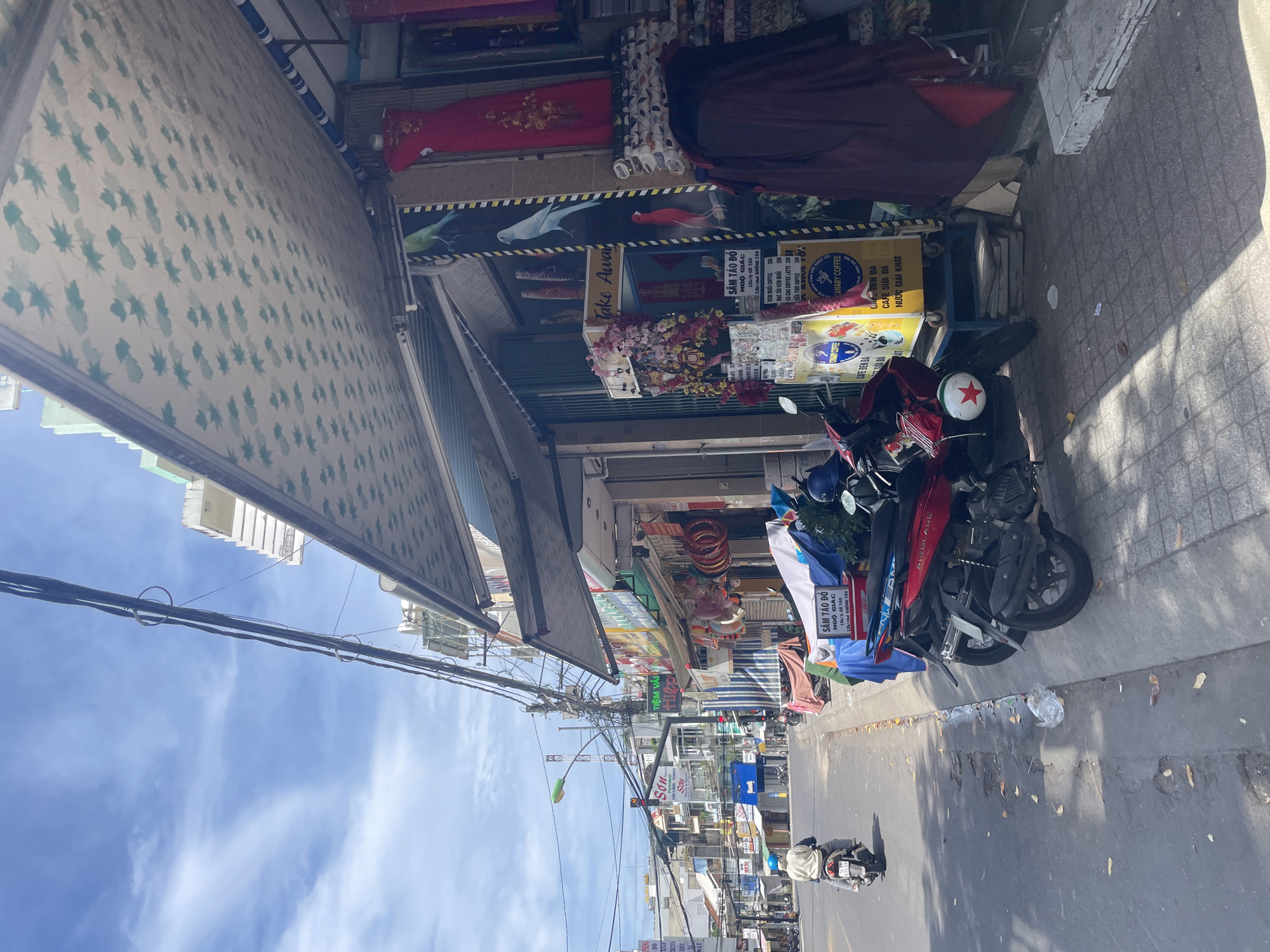 The image shows a street scene in Nha Trang, Vietnam, with a focus on a storefront and the road in front of it. A blue sky with scattered clouds is visible above. On the right side of the image, a storefront displays various items, including clothing on mannequins, fabric rolls, and decorative items like red rings. A prominent awning with a repeating pattern extends over the storefront.
In front of the storefront, two motorcycles are parked. One is a red motorcycle with a white star decal on its front, and the other is a darker colored motorcycle. A person wearing a light-colored jacket and blue helmet is riding a motorcycle away from the camera down the paved road on the left side of the image. The road has some potholes and scattered leaves.
There are numerous electrical wires strung across the street, supported by poles. Various signs with Vietnamese text are visible on the buildings along the street. A yellow food cart with "CANARY COFFEE" written on it is also present near the storefront. The overall impression is that of a bustling street in a Vietnamese city, likely a commercial area with shops and ongoing street life.