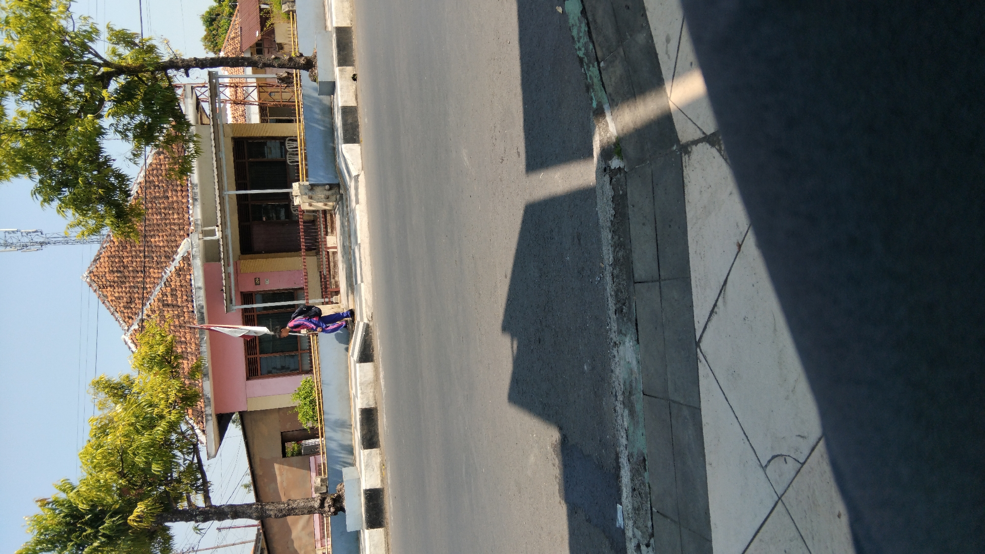 The image depicts a bright, sunny day in the city of Batang, Indonesia. A pink and yellow building with a tiled roof stands on the right side of the frame, with trees flanking it on either side. A person in a blue tracksuit and backpack walks on the sidewalk in front of the building. The street in front of the sidewalk is paved with asphalt, and a shadow of the building is cast across it. The sidewalk is made of concrete and tiles. A cell tower is visible in the background behind the building. The overall scene suggests a residential or public building in a suburban area.