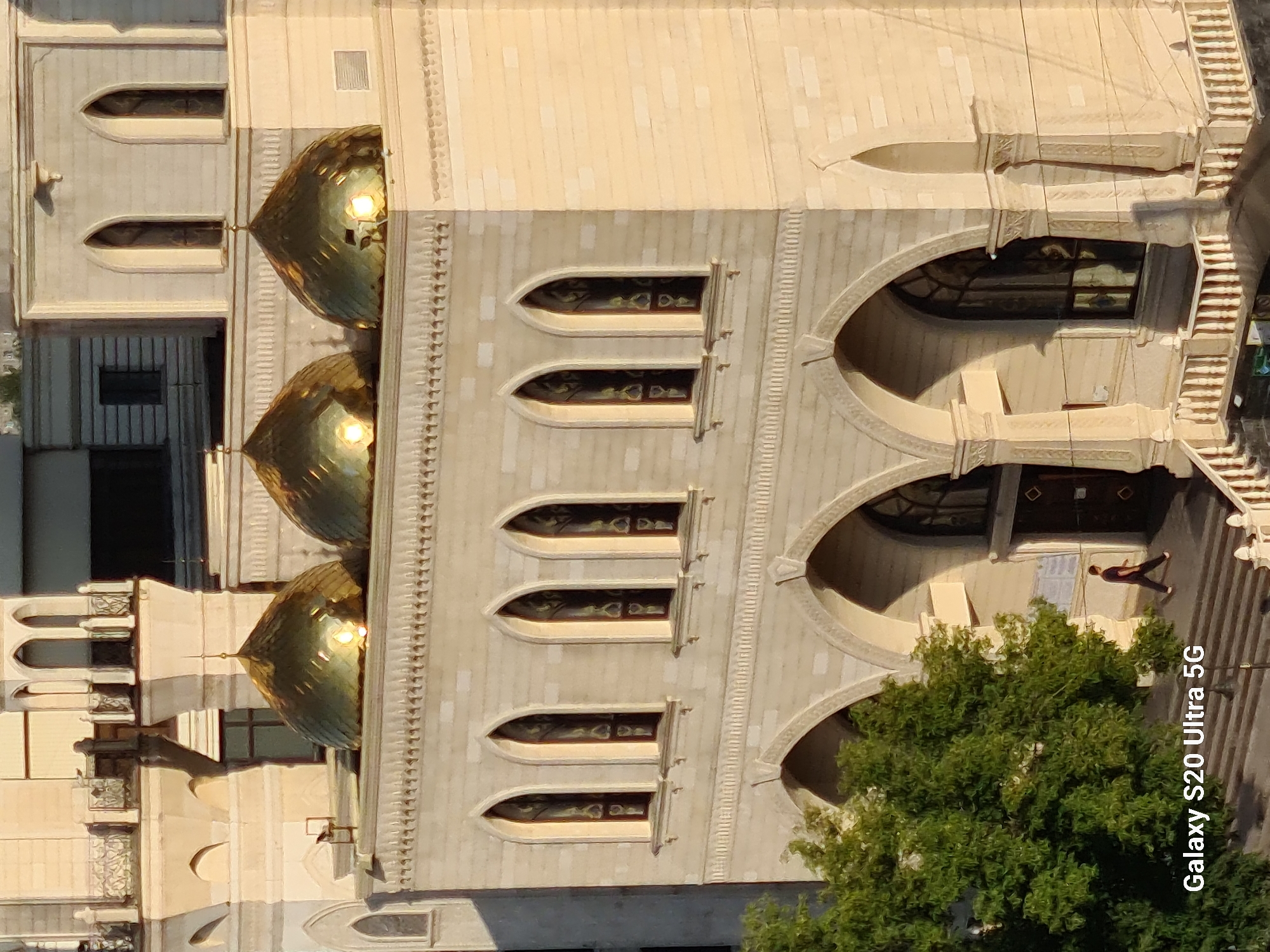 The image shows an exterior view of the Central Mosque of Almaty, Kazakhstan, captured from a high vantage point. The mosque features a light-colored stone facade with multiple arched windows and a series of golden domes. In the foreground, to the left, a large green tree is visible. A single person, dressed in dark clothing, is walking up a set of stairs towards the mosque's entrance. The lighting suggests it is either early morning or late afternoon, with the sun casting long shadows. The "Galaxy S20 Ultra 5G" watermark is present in the lower left corner of the image.