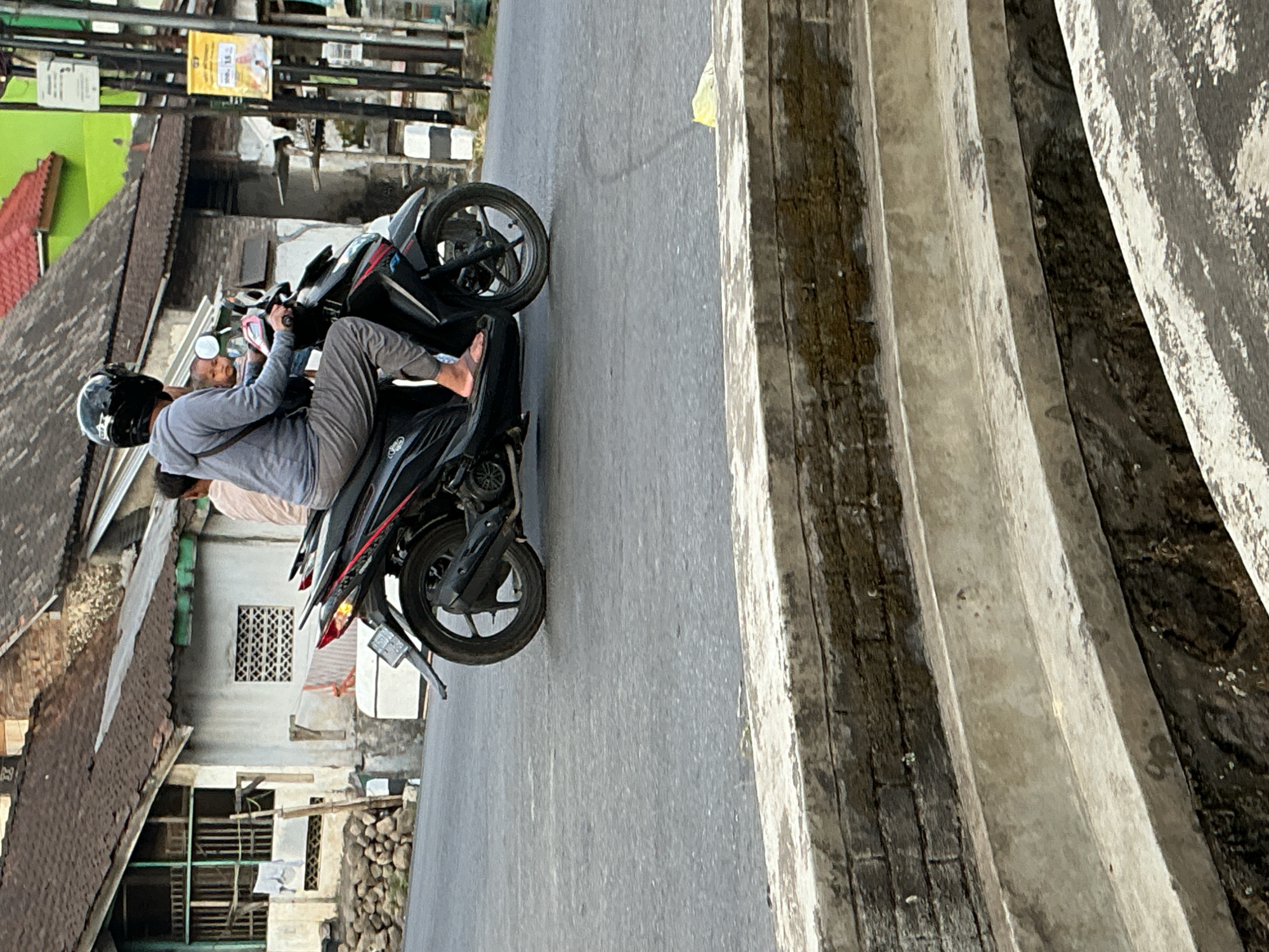 In Kebonrejo, Indonesia, a person wearing a black helmet and a grey hoodie rides a black motorcycle. They are wearing grey pants and flip-flops. A child is seated in front of the rider. The motorcycle is on a paved road. In the background, there are houses with tiled roofs, some with visible laundry hanging. A yellow sign is attached to a pole on the right side of the image. In the foreground, there is a concrete structure with several steps.