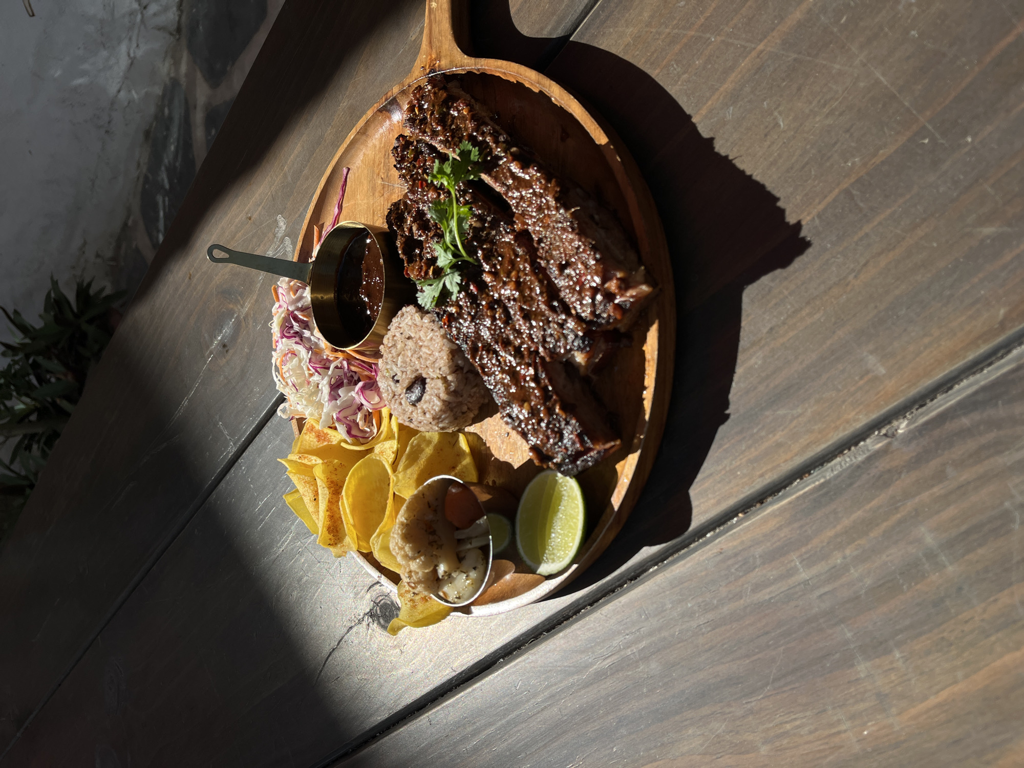 A wooden platter of food sits on a wooden table. The platter includes meat, rice, slaw, plantain chips, pickled vegetables, and lime. A small metal saucepan of dark sauce also rests on the platter. The scene is lit by bright sunlight. Đà Nẵng, Vietnam is not mentioned, nor is its presence implied.