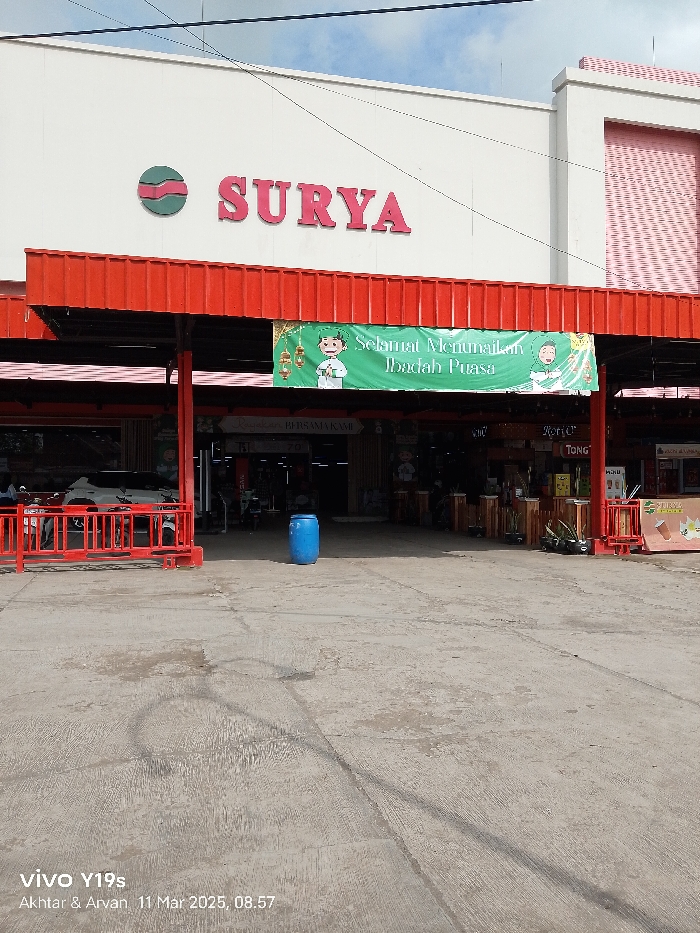 The image shows the front entrance of a store named "SURYA" in Jatibarang, Indonesia. It is located at coordinates -6.467 latitude and 108.298 longitude. The store has a red awning over the entrance and a sign that says "Selamat Menyambut Bulan Puasa" which means "Happy to welcome the fasting month". There is a red metal barrier across the entrance and a blue plastic barrel sitting on the concrete pavement. The entrance is empty, and the sun is shining brightly. There are multiple windows and a doorway behind the awning. The ground is concrete and is streaked with tire marks. There is a second awning and signage on the roof of the building to the left of the entrance. The image was captured by a Vivo Y19s on March 11, 2023 at 08:57.