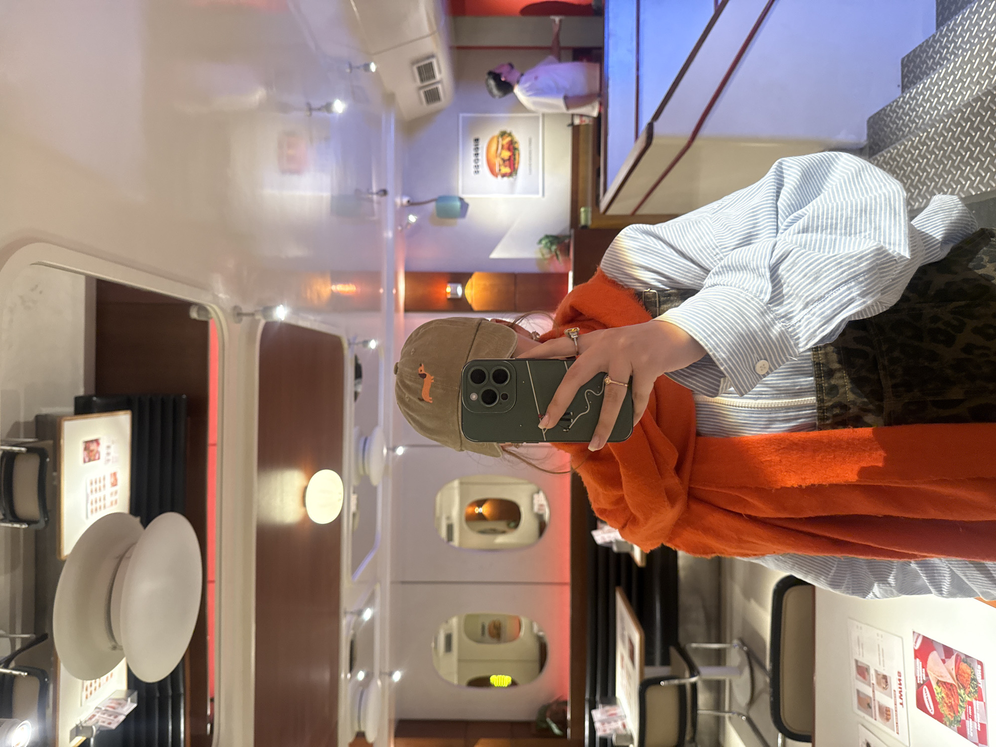 A person is taking a selfie in a brightly lit interior. The interior appears to be a cafe or restaurant with a futuristic or retro design. The person is wearing a light blue and white striped shirt, a leopard print jacket, and has an orange scarf draped around their shoulders. They are also wearing a tan baseball cap with an orange fox logo. They are holding a green smartphone up to take the picture. The background shows the interior of the restaurant, including white walls with porthole-like windows, lighting fixtures, tables, and posters. In the background, there is a poster for a burger and a person wearing a white shirt. A sign with menu options is on the table. The city of Yerevan, Armenia is not evident from the image.