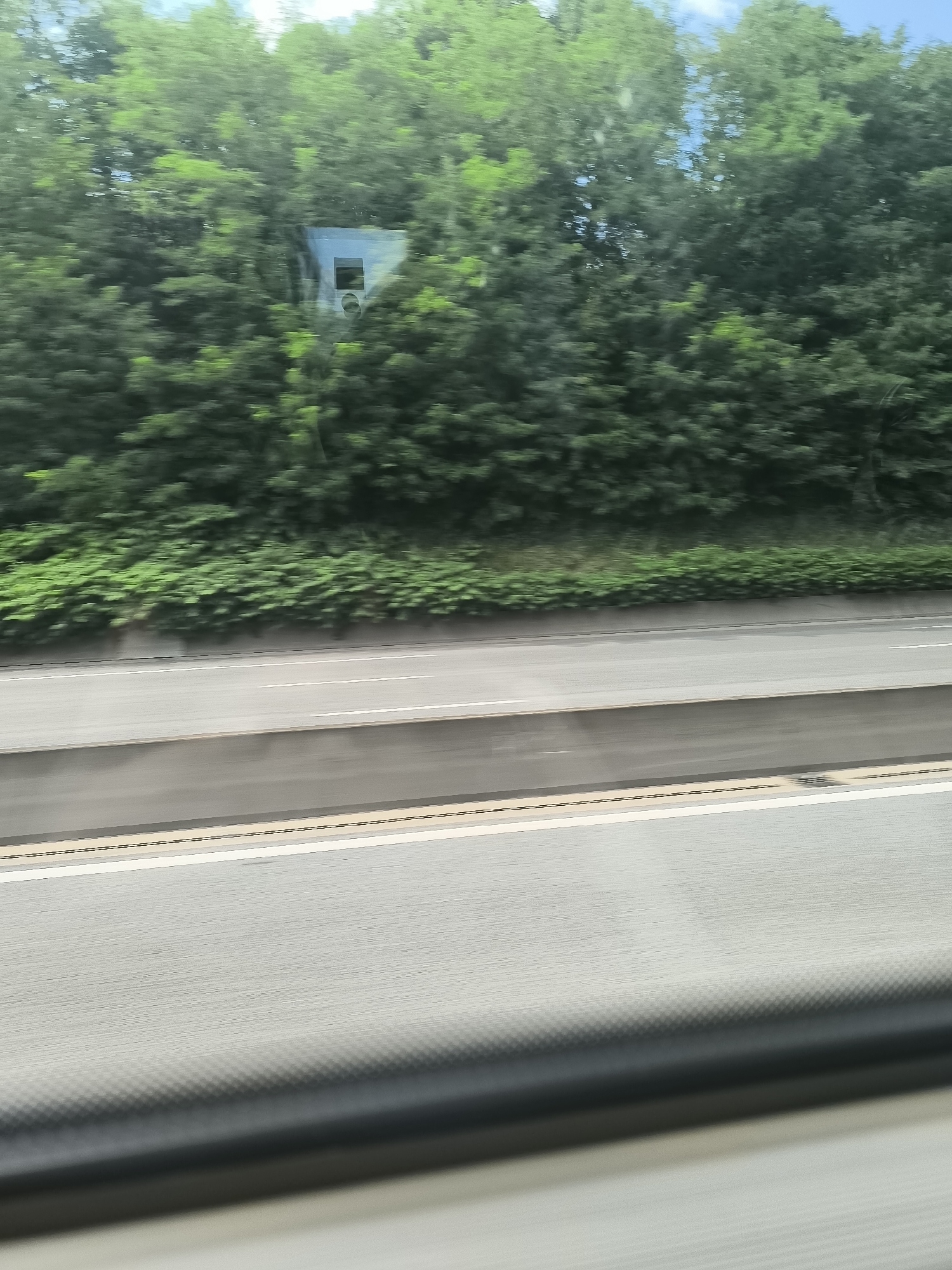The image shows a view from inside a moving vehicle traveling alongside a highway. The highway has multiple lanes and is bordered by dense green vegetation. Partially obscured by the foliage is a small structure, possibly a utility box or a camera housing. The lower portion of the frame is dominated by the interior of the vehicle, with a portion of the dashboard visible.
The location is not discernible based on the provided image. Saint-Jean-Bonnefonds, France, cannot be confirmed as the specific location without additional context or visual cues.