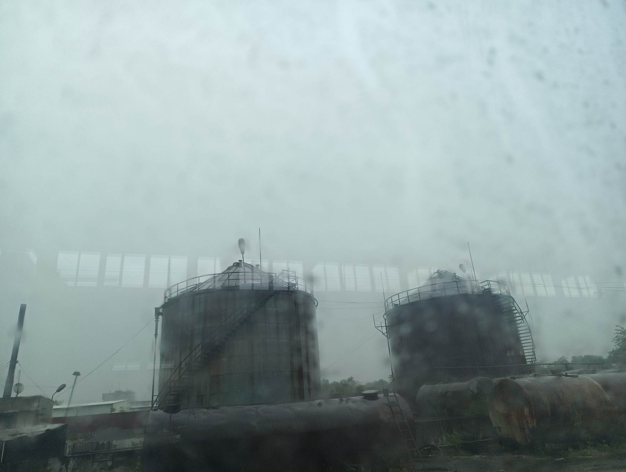 The image captures a hazy industrial scene featuring two prominent, large cylindrical storage tanks, possibly silos, in the midground. The photograph appears to be taken through a wet or fogged window, as indicated by visible water droplets and streaks, which contribute to the soft focus and muted color palette.
Both large tanks are weathered, with dark, metallic surfaces, and each is equipped with external ladders and railings leading to their conical roofs. A plume, possibly steam or mist, is visible rising from the top of the left tank. In the foreground and between the larger structures, several smaller, horizontal cylindrical tanks are scattered on the ground, appearing aged and somewhat rusted. To the far left, a tall, slender pole or chimney stands partially visible.
A large, low-rise industrial building with distinct, rectangular window frames or structural elements spans the background, further blurred by the atmospheric conditions. Overhead lines or cables are also faintly discernible. Patches of sparse, dark vegetation are visible around the base of the tanks and in the foreground.
The overall setting is an industrial complex or storage facility, suggesting a manufacturing or processing environment. The prevailing weather is extremely hazy, foggy, or misty, creating a desolate and quiet atmosphere. No people are visible within the scene, and there is no discernible text. The time of day appears to be daytime, heavily obscured by the thick atmospheric conditions.