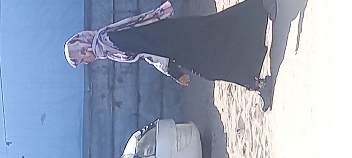 A woman wearing a headscarf and a dark, full-length garment walks on a sandy or light-colored surface. Her shadow is visible. A vehicle, possibly a car, is partially seen to the side of her. The background includes a blue wall or structure next to a weathered, wooden-like partition.