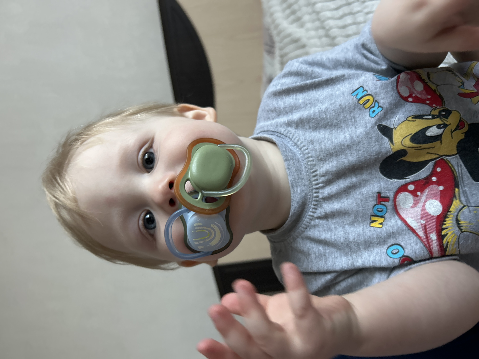 A fair-skinned baby with light hair and blue eyes is captured in a close-up, medium-shot photograph, rotated 90 degrees counter-clockwise. The baby is looking directly at the viewer with a neutral, slightly curious expression. Two pacifiers are in the baby's mouth; the upper one is green and orange, and the lower one is blue and light brown with a faint pattern.
The baby is wearing a gray long-sleeved t-shirt featuring colorful graphics. Visible designs include Disney's Mickey Mouse and Minnie Mouse characters, along with red and white spotted mushrooms. Text printed on the shirt includes "RUN" in blue and yellow letters, and "NOT" in yellow and blue letters. The baby's hands are visible in the foreground, slightly blurred, suggesting movement or an outstretched gesture.
The background is simple and out of focus, consisting of light-colored walls or furniture, with a darker vertical element (possibly a piece of furniture or doorframe) on the left side of the frame. The overall setting appears to be indoors. The image provides no specific cues for the time of day or weather. The location context is Chaikovsky, Russia.