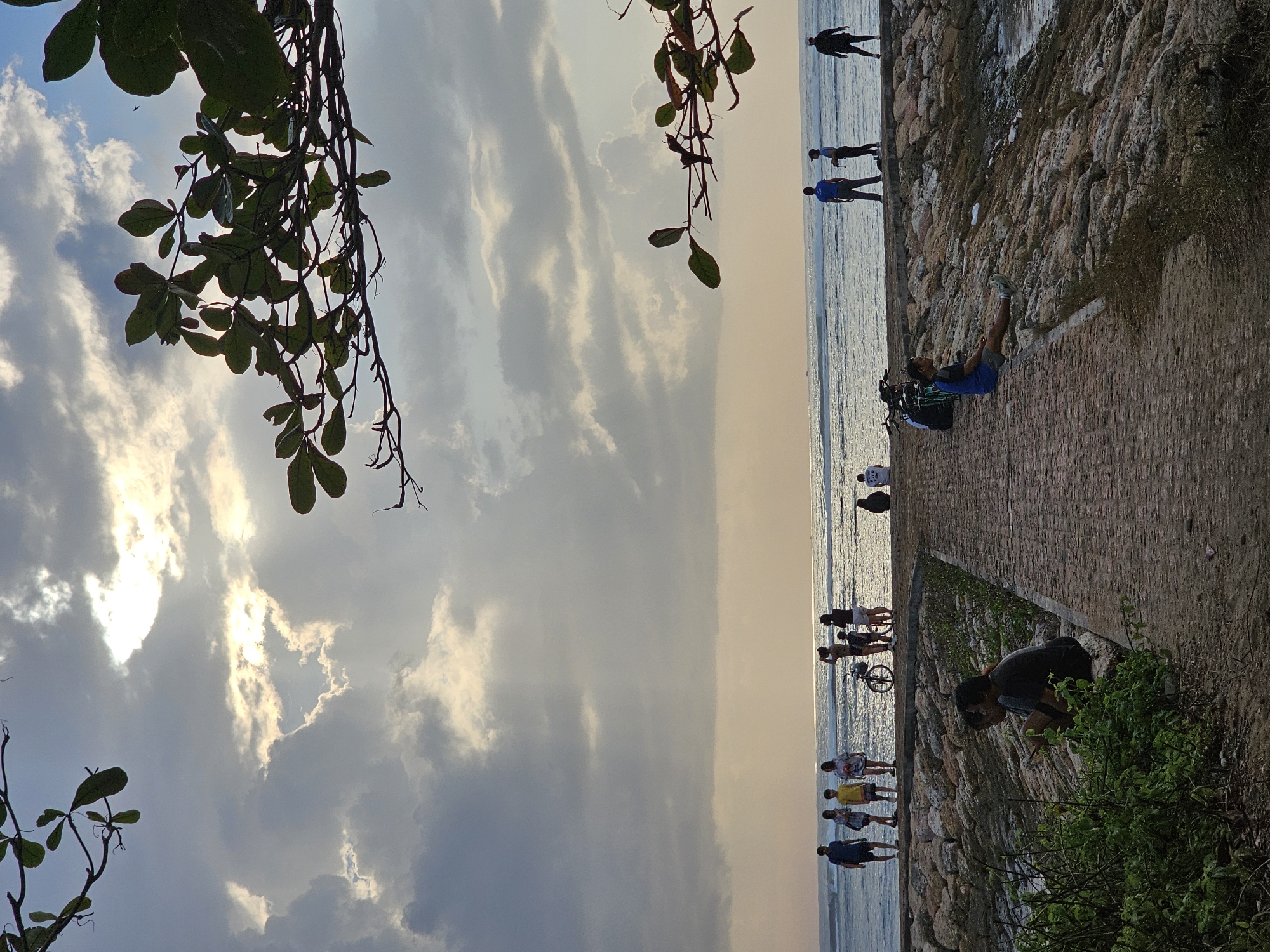 The image depicts a tranquil scene at a beach in Denpasar, Indonesia, likely during sunset. A pathway made of cobblestones leads towards a rocky embankment overlooking the ocean. Several individuals are scattered across the scene. On the pathway, two people are seated near lush green foliage. Further along the embankment, a group of people are standing and observing the sea, some with bicycles. In the distance, a single person is seated on the embankment. The ocean is calm with gentle waves, and the sky is filled with dramatic clouds partially illuminated by the setting sun. Lush green leaves from nearby trees frame the top of the image.