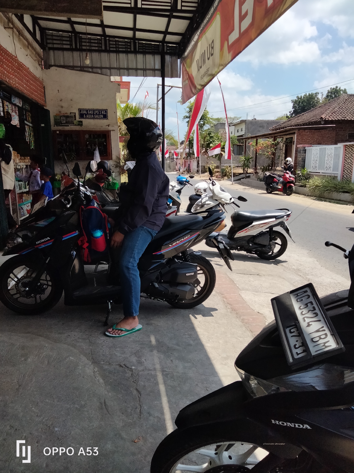 In the city of Kelutan, Indonesia, a person wearing a black helmet and a dark jacket is seated on a black Honda scooter. The scooter has a red and blue stripe and a backpack with red and blue accents is attached to its side. The person is wearing blue jeans and green flip-flops. The scooter is parked on a sidewalk next to a small shop that sells gas cylinders and bottled water, indicated by a sign that reads "JUAL GAS LPG 3 KG & AQUA GALON". Indonesian flags are visible in the background. Several other scooters are parked nearby. The scene appears to be a street-side commercial area with buildings and trees lining the road.