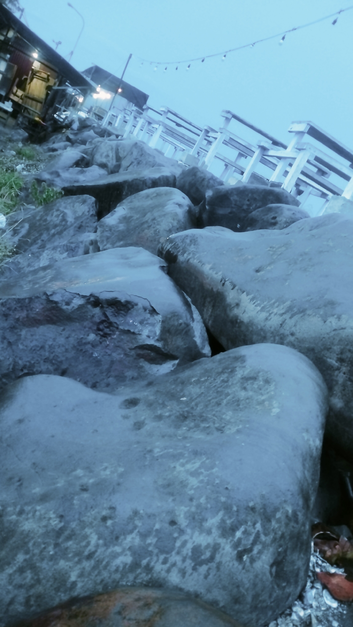 This image captures a tranquil twilight or early evening scene, likely along a coastal area in Lhokseumawe, Indonesia. The foreground is dominated by large, wet, irregularly shaped rocks, appearing dark blue-gray, possibly functioning as a breakwater or sea wall. Some green vegetation and scattered debris are visible amongst the rocks on the left.
In the mid-ground, a row of white wooden benches or railings lines the embankment, suggesting a promenade or seating area overlooking water. Behind these, several structures resembling small buildings or stalls, possibly cafes or eateries, are dimly lit, their warm internal lights contrasting with the cool blue ambiance. A lamppost is partially visible towards the center-left. Above the structures and benches, a string of decorative bulbs or fairy lights stretches across the sky, further indicating the evening hour.
The sky is a uniform, pale blue-gray, suggesting an overcast evening or twilight conditions. The wet appearance of the rocks could imply recent rain or the proximity to the sea. No people or specific activities are depicted, giving the scene a calm and quiet atmosphere. There is no visible text in the image.