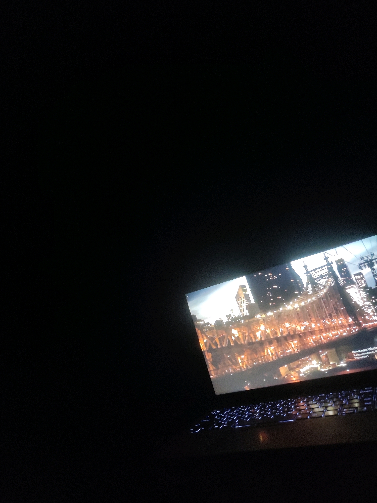 A laptop is placed on a dark surface, angled upwards. The screen displays a nighttime cityscape with a bridge illuminated by numerous lights. The keyboard of the laptop is visible in the foreground and is backlit with blue light. The background is entirely black, suggesting the image was taken in a dark environment. There are no people or other objects visible in the scene, nor any indications of the location being Burgoberbach, Germany.