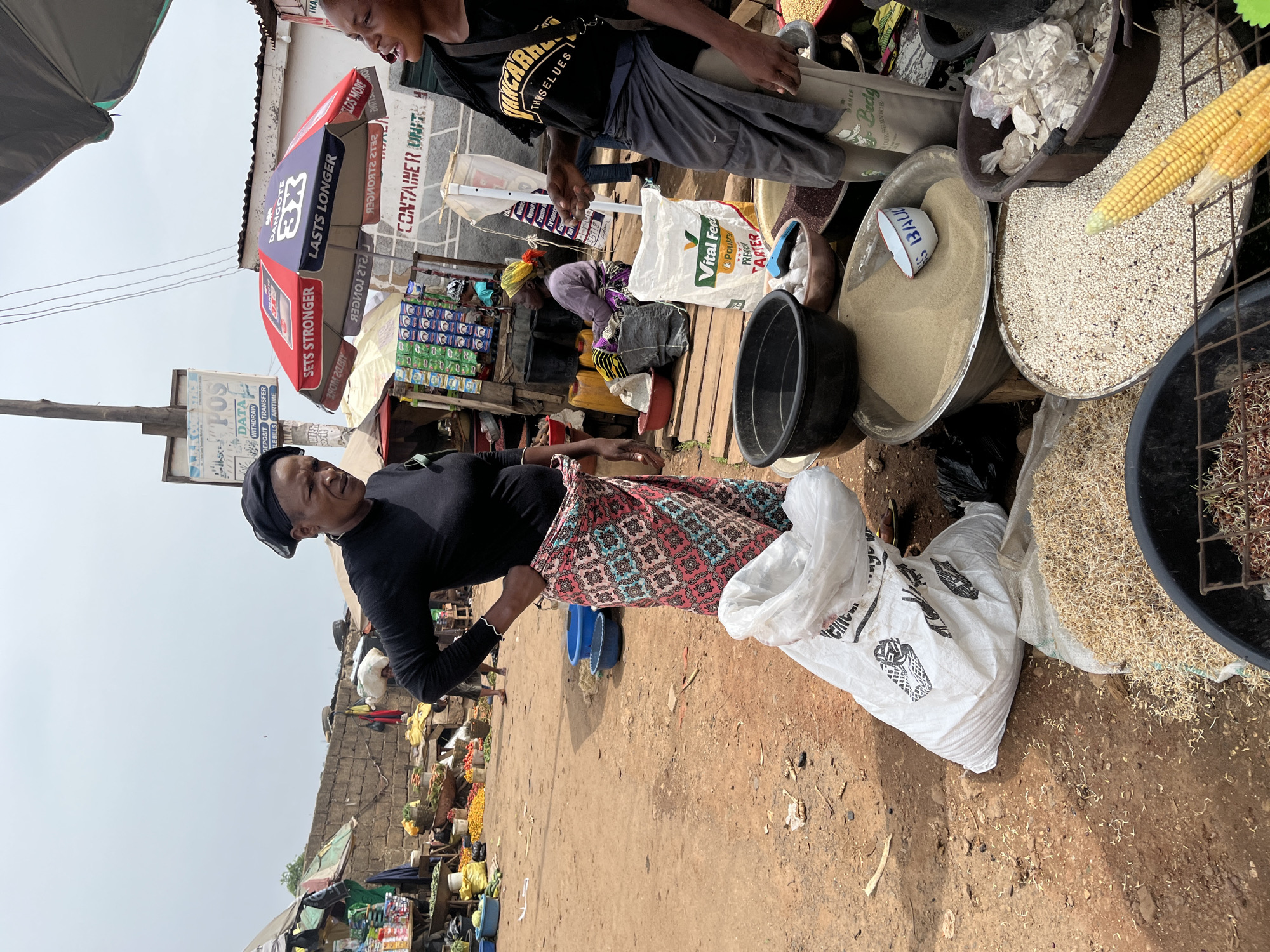 The image depicts an outdoor market scene in Rafin Par, Nigeria. Two women are the primary subjects. The woman on the left, wearing a black long-sleeved shirt and a patterned skirt, stands with her hands on her hips, facing slightly to the right. She has a headscarf covering her hair. To her right, a younger woman, wearing a black t-shirt with yellow lettering and a grey skirt or wrap, is speaking and gesturing with her right hand. She is partially obscured by a white bag to her left.
The environment is dusty and unpaved. Stalls and tables are visible in the background, laden with goods. To the left, there are stacks of what appear to be food items, including vegetables and yellow fruits. Further back, a brick wall and various structures suggest the presence of permanent market buildings. An umbrella with "DANGOTE" branding is prominently displayed. In the foreground, a white sack of grain is placed on the ground, and several large bowls contain various foodstuffs, including a fine sand-like substance, white grains, and what appear to be sprouts. A partially eaten ear of corn is also visible in one of the bowls. The overall impression is a bustling, open-air marketplace.