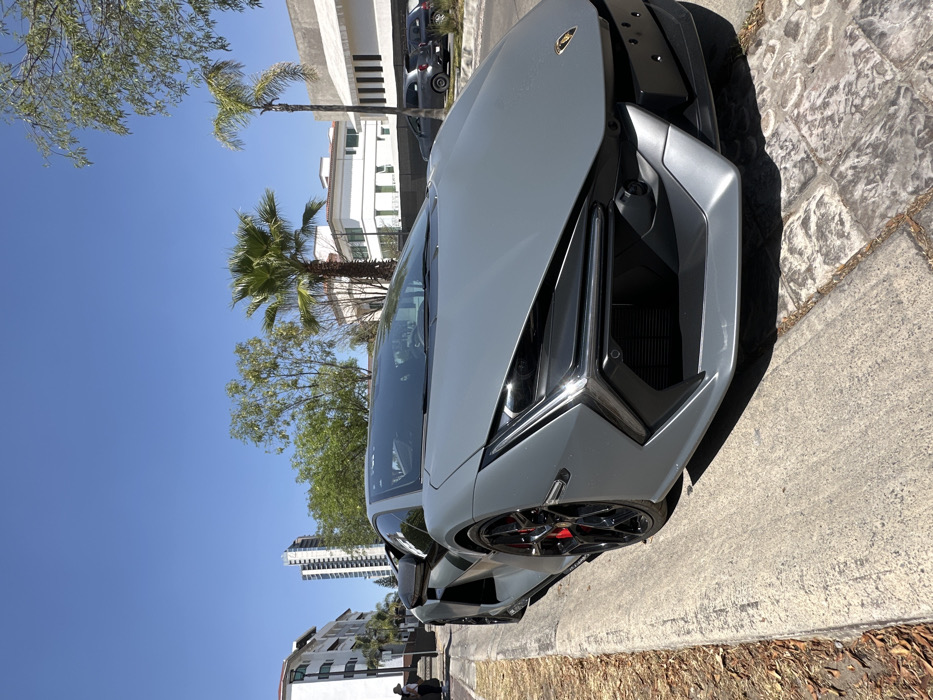 A matte grey Lamborghini Revuelto is prominently parked on the right side of a concrete curb, adjacent to a paved street surface, under a clear blue sky in San Bernardino Tlaxcalancingo, Mexico. The vehicle's front right quarter is angled towards the camera, displaying its sharp angular headlights with Y-shaped daytime running lights, a distinct front grille, black multi-spoke wheels, and red brake calipers. A black, empty license plate bracket is affixed to the front bumper. In the background, modern white buildings with contemporary architectural styles line the street. A tall, slender skyscraper is visible in the mid-distance to the left. Green trees and several tall palm trees are interspersed throughout the scene. To the far left, a partially visible person wearing a white shirt and dark lower garments stands near one of the buildings. Further down the street on the right, other vehicles, including a dark grey hatchback, are parked. The bright sunlight casts distinct shadows, indicating daytime conditions.