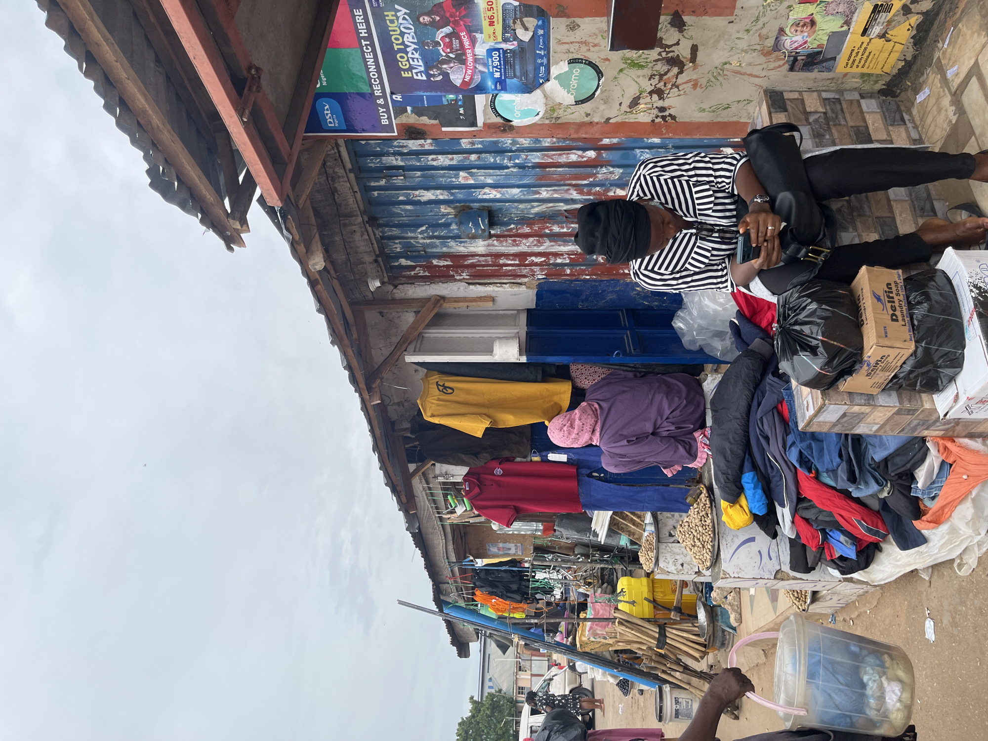 A woman in a black and white striped shirt and black pants sits on a low wall, looking down at her phone. She wears a black headscarf. To her left are stacks of boxes and black plastic bags. To her right is a corrugated metal wall covered with posters and advertisements, some of which are torn. In the background, a market stall displays clothing and other goods. Several other people are visible in the distance, including a woman in a purple outfit and headscarf, and someone carrying a clear plastic bucket. The market appears to be outdoors, with a cloudy sky overhead. This scene is located in Gura Top, Nigeria.