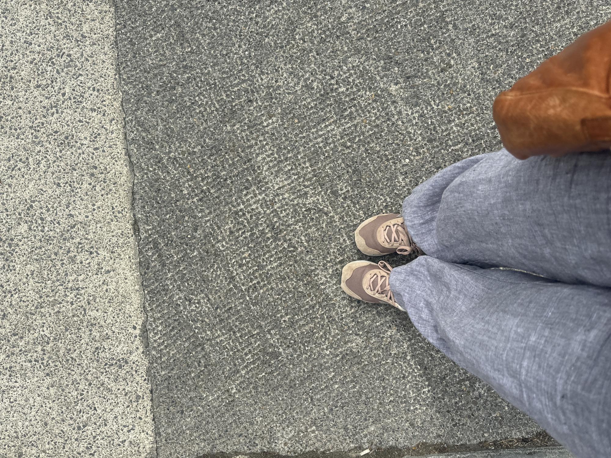 A person stands on a textured gray pavement in Waterford, Ireland, their feet clad in tan and pink sneakers and loose-fitting grey trousers. They are holding a brown leather handbag. The pavement is divided into two distinct textures, with a lighter, coarser aggregate material meeting a darker, finer aggregate material.