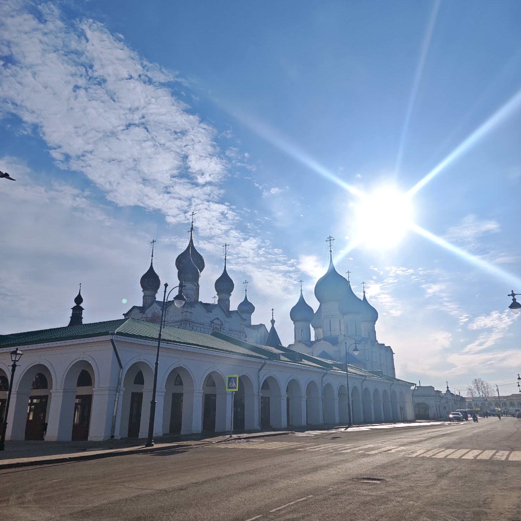 The image displays a sunny street scene in Rostov, Russia, featuring the Market Arcades and a portion of the Rostov Kremlin complex. The primary subject is a long, white, single-story building with a green sloped roof, characterized by a series of approximately twelve evenly spaced arched openings along its facade, extending from the lower left towards the mid-right. A yellow pedestrian crossing sign is positioned on a pole in front of this building. Behind and above this structure, multiple white Russian Orthodox churches are visible, featuring numerous dark, light-blue, or grey onion domes, each topped with a cross. The domes vary in size and height, with the largest and most prominent appearing on the right side of the main church structure.
The sky is bright blue with scattered white cumulus clouds, particularly concentrated on the left side of the frame. A strong sun is positioned in the upper right quadrant, emitting a prominent lens flare with visible rays.
In the foreground, an asphalt road occupies the lower portion of the image, featuring white "zebra crossing" markings that extend across its width. A narrow sidewalk runs parallel to the arcade building on the left. A black, ornate streetlight stands on the left side of the road, with a portion of another visible on the far right.
On the road in the distance, towards the right, several vehicles are visible, including what appears to be a light-colored sedan and a darker SUV. A few indistinct human figures are also present on the far right side of the road, suggesting pedestrian movement. A small, dark bird is captured in flight in the upper left corner. The distant background on the right side of the street shows additional light-colored buildings and some bare trees, indicative of a dormant season.