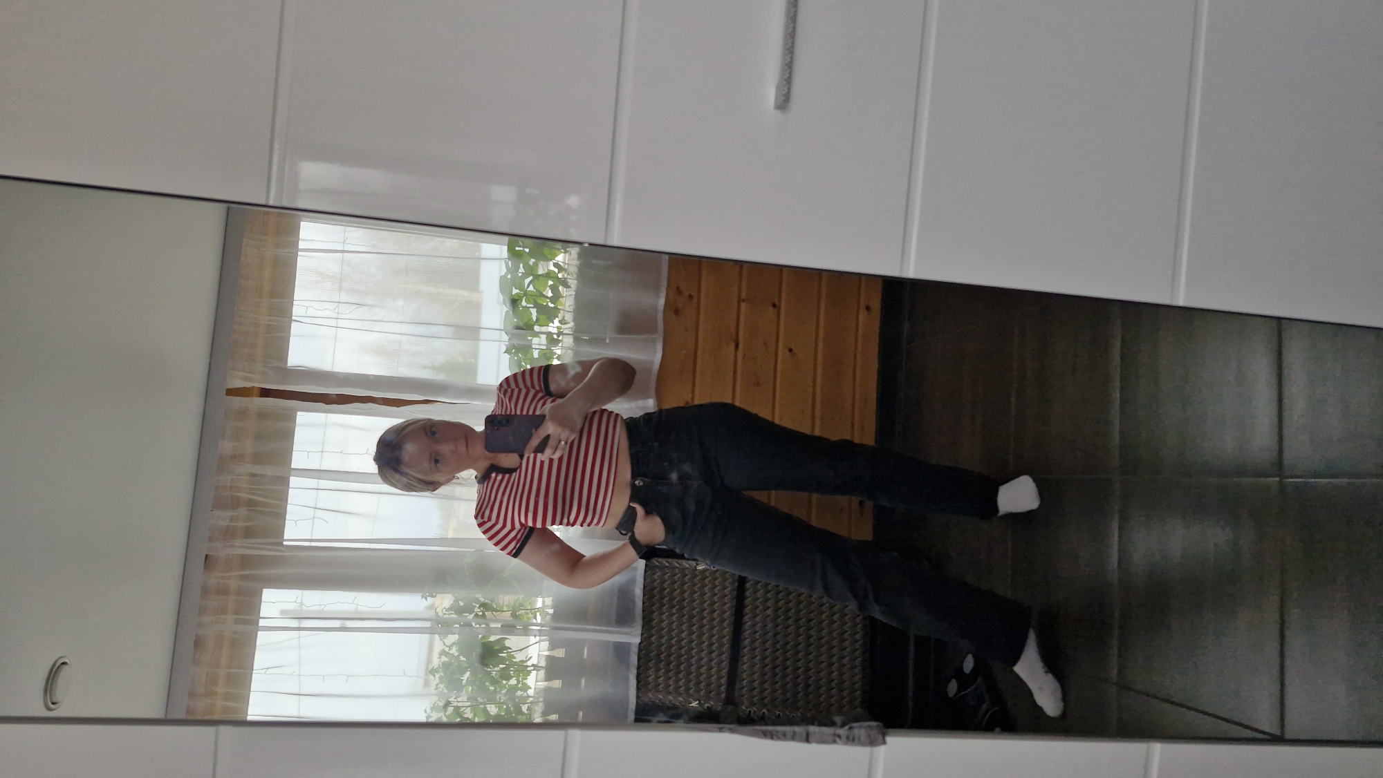 A young, light-skinned individual with short, blond hair is reflected in a large mirror. The individual is wearing a red and white striped crop top, dark denim jeans, and white socks. They are holding a smartphone, presumably taking a photo of themselves.
The background reflected in the mirror includes a window with light-colored curtains, a wood-paneled wall, a plant visible through the window, and a dark tiled floor. The mirror itself is set against a white wall with vertical paneling. Based on the reflection, the location is inside a room in an environment with access to natural light and elements of domestic interior design. No landmarks are visible that confirm the location as Otchor, Russia.