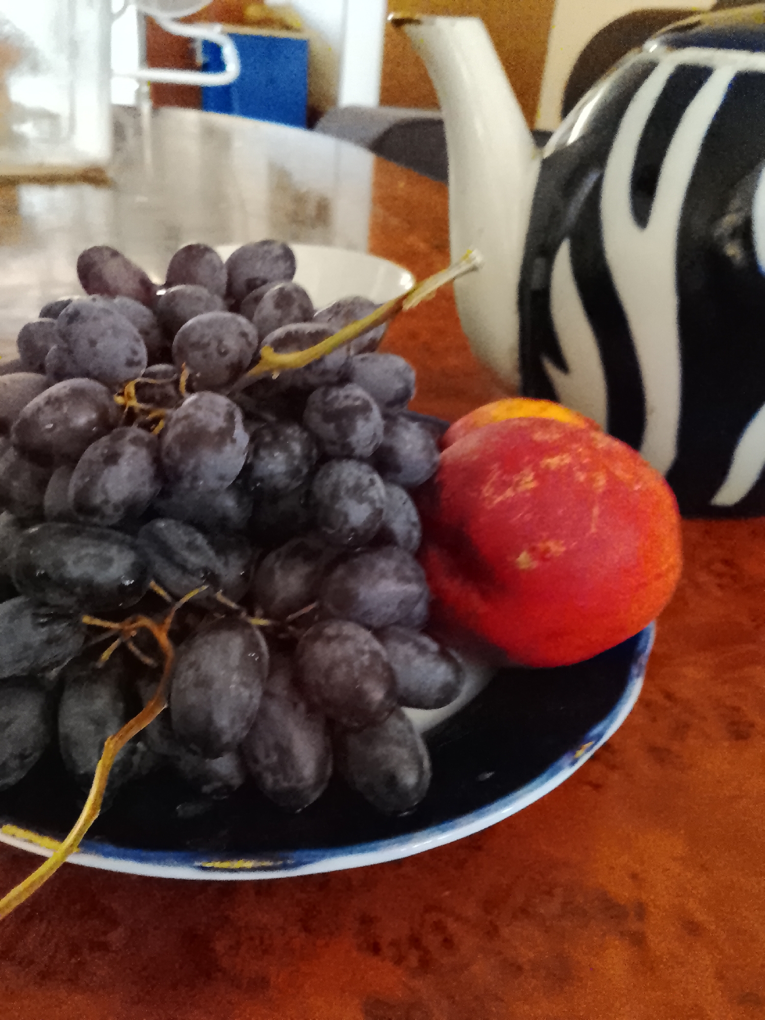 A close-up shot shows a dark plate of black grapes and a red nectarine on a brown, wood-grain table. To the right of the plate, a white teapot with black zebra stripes is visible. The background is blurred, suggesting an indoor setting with hints of light-colored furniture and possibly some blue objects. The lighting is soft and natural. There are no people or animals in the frame, and no specific identifiable landmarks of Kasri Arifan, Uzbekistan are present.