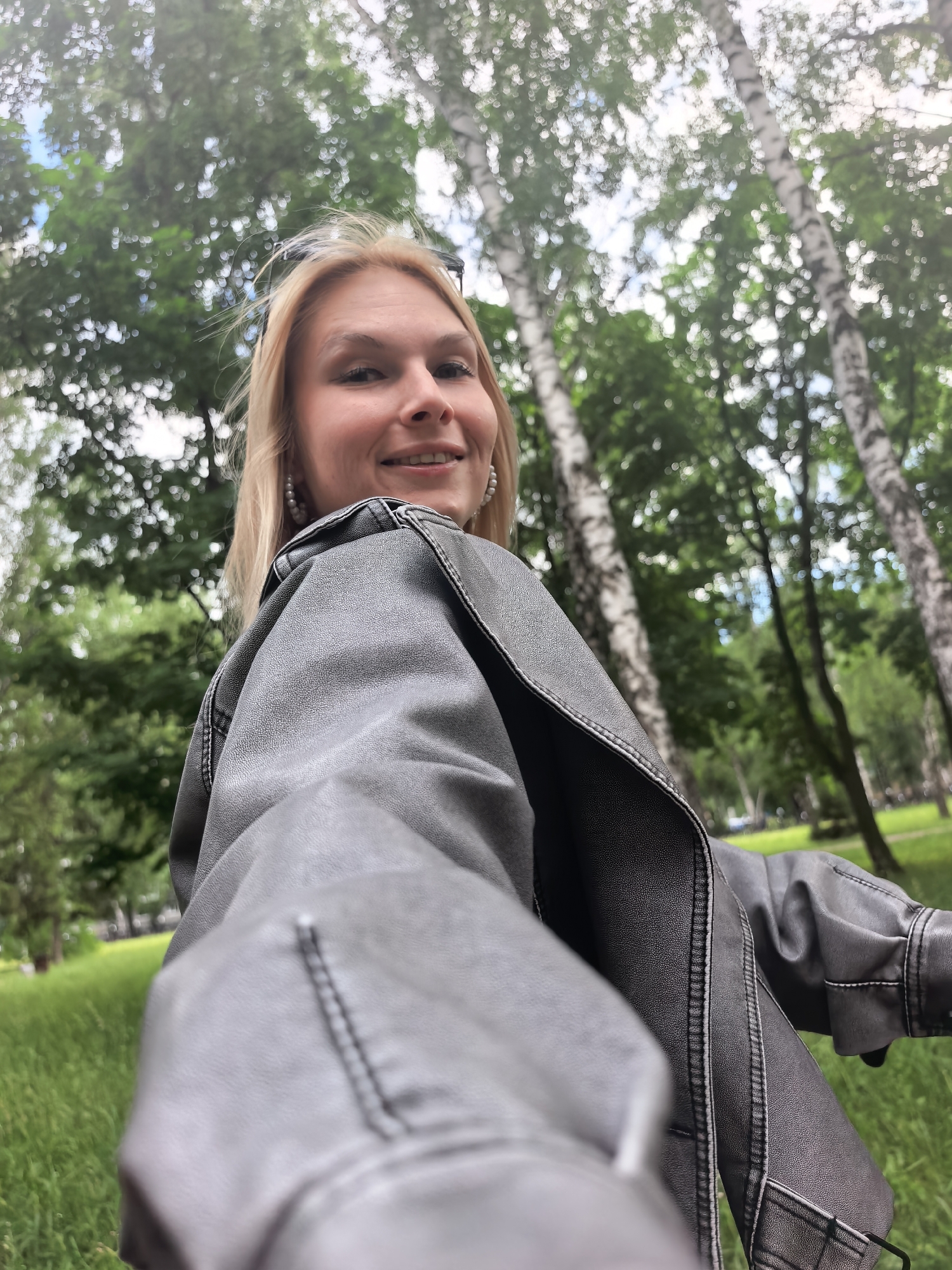 A woman with blonde hair is taking a selfie outdoors. She is wearing a grey leather jacket and earrings. Behind her is a park with green grass and trees, including white-barked birch trees. Based on visual markers such as the birch trees, the location is likely in a park in Russia. The city is Sterlitamak, Russia.