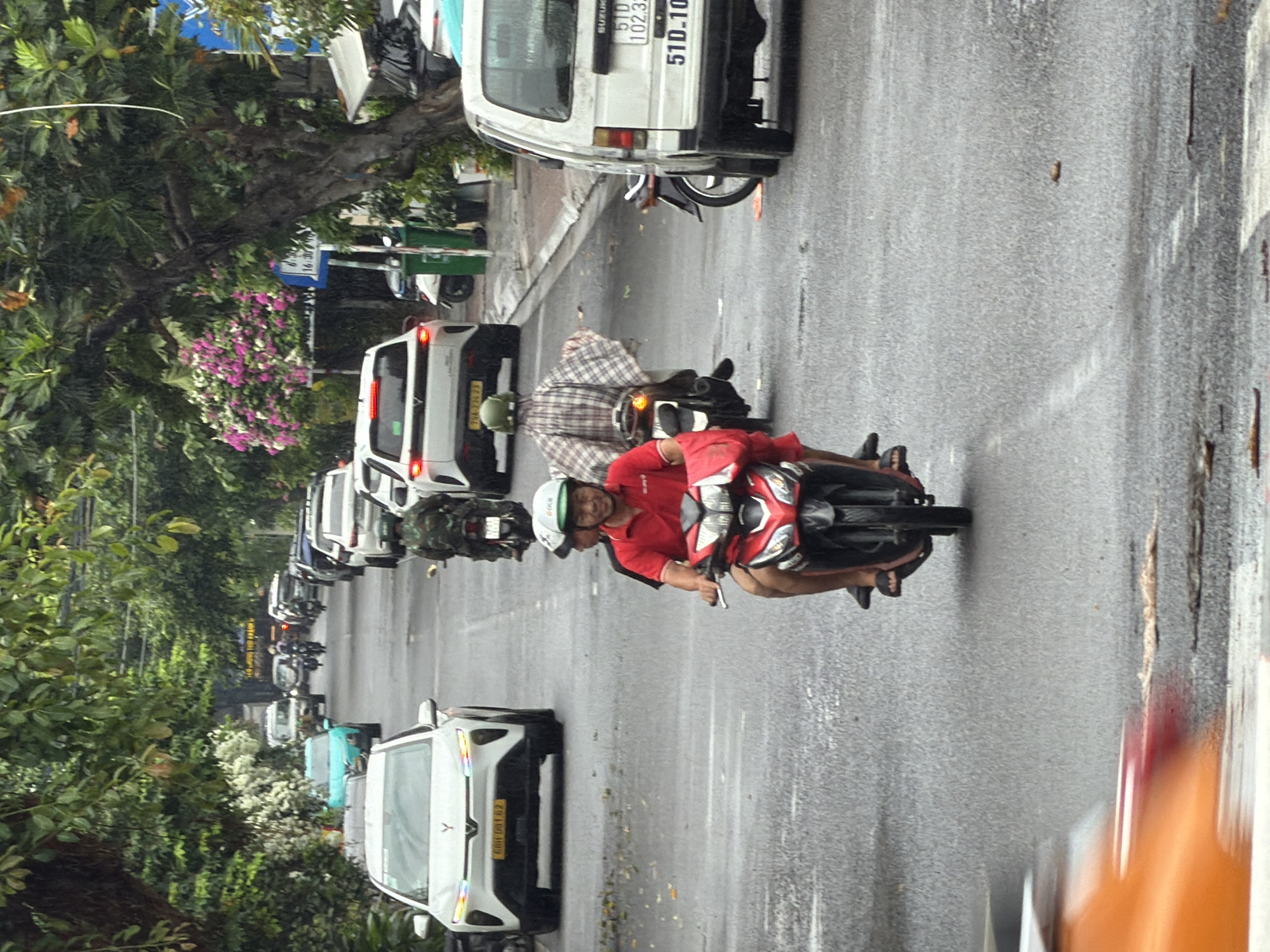 A street scene in Nha Trang, Vietnam, shows a wet asphalt road with several vehicles and pedestrians. In the foreground, a person wearing a red shirt and a green helmet rides a red scooter. Behind them, another person wears a plaid shirt and a green helmet, also on a scooter. To the right, a white Suzuki van is parked, displaying a license plate with the characters "51D-102.33". Further down the road, a white car with a yellow license plate is visible, along with other cars and motorcycles. Lush green trees and flowering bushes line the sides of the road, suggesting a tropical environment. The road appears slick, indicating recent rain.