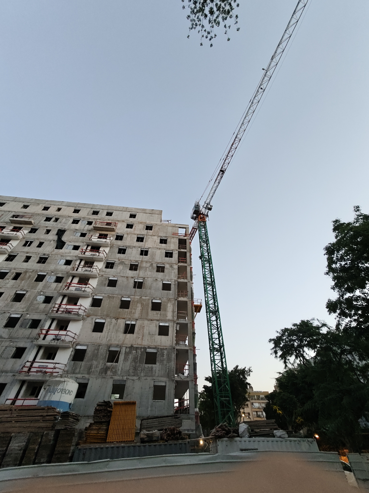 The image captures an unfinished concrete building under construction, likely in Petah Tikva, Israel, given the context. A tall green tower crane dominates the right side of the frame, its boom extending diagonally upwards into a clear sky. The building itself is several stories high, with numerous windows and balconies, some with red railings. Construction materials, including stacks of wood and what appear to be concrete forms, are visible at the base of the building. Trees, some with lush green foliage, surround the construction site. The scene suggests an active construction environment during daylight hours.