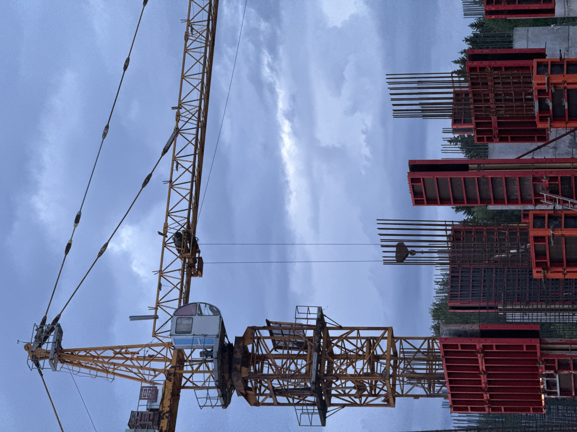 The image depicts a construction site in Izhevsk, Russia. A yellow tower crane dominates the left side of the frame, contrasted against a cloudy sky. The crane is oriented vertically. To the right, forms and reinforced steel structures indicate the preparation for concrete pouring, possibly for a building foundation or a wall. Trees are visible in the background. The scene suggests ongoing construction activity. No people are visible.