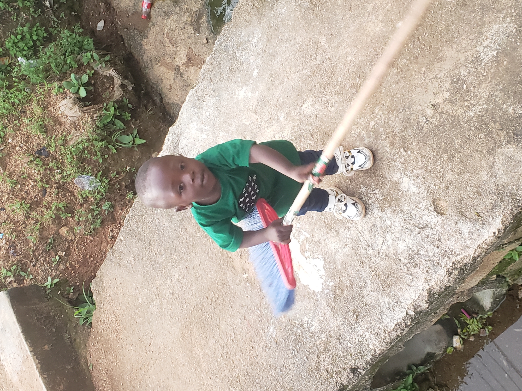 A young boy, appearing to be between two and four years old, stands on a concrete pathway in what is identified as Jos, Nigeria. He is wearing a green t-shirt with a graphic design on the front and dark pants. On his feet are white sneakers with black and gold accents. The boy is holding a broom with a red and blue brush head and a light-colored wooden handle, positioned as if he is sweeping. The pathway appears to be elevated, with a slope leading down to a drain or ditch on the left side of the image. This ditch contains murky water, green vegetation, and some debris, including a pink object. To the right of the pathway, there is a dirt embankment covered in sparse green plants and some litter, including a red plastic bottle. The overall environment suggests an outdoor, possibly semi-urban or rural, setting.