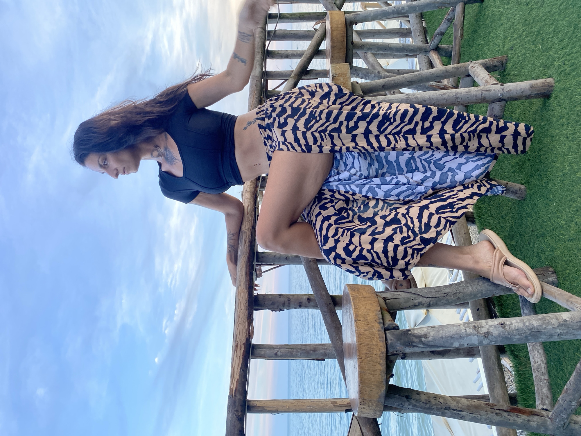 An adult female with long, dark hair, wearing a black crop top and a long, flowing skirt with a tiger-stripe pattern, is seated on a rustic wooden stool. She is looking to her right, with her left leg crossed over her right. She has tattoos on her neck, arms, and torso. The stool is made of rough-hewn wood and is positioned on a green, artificial turf surface. Behind her, a wooden railing overlooks a body of water, likely the sea, with a pale blue sky above. The scene appears to be an outdoor seating area near a waterfront. The location is identified as Phu Thuy Ward, Vietnam.