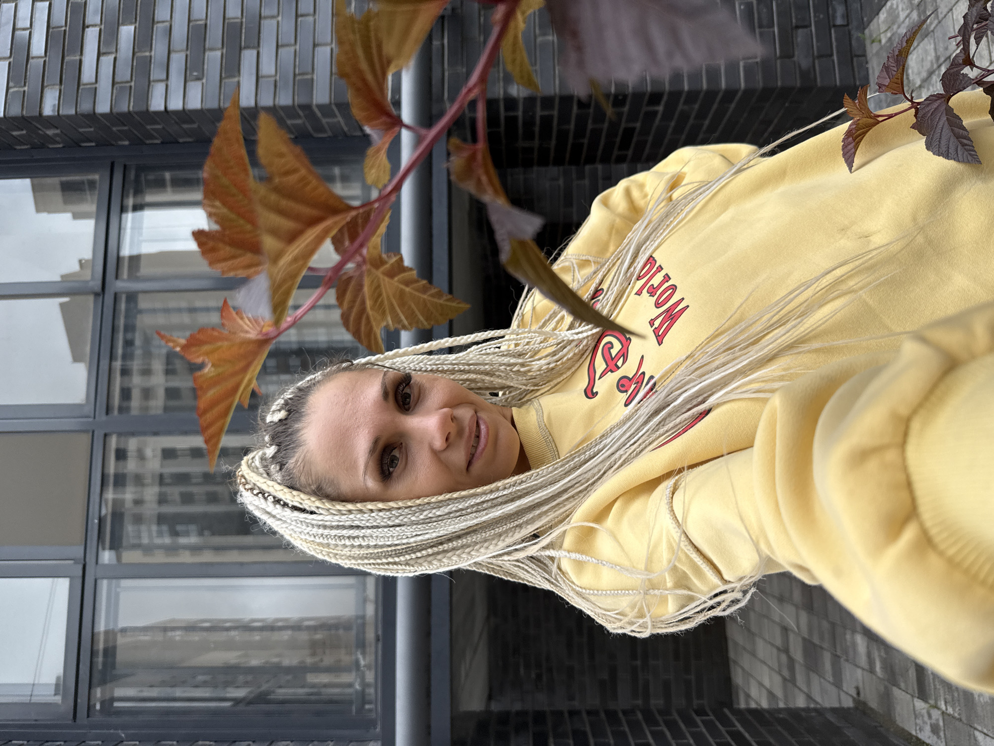 A blonde woman with many thin, light-colored braids is taking a selfie outdoors in front of a modern building with dark brickwork and large glass windows. She is wearing a yellow sweatshirt with red lettering that partially reads "World". A branch with reddish-brown leaves is in the foreground on the right side of the image, obscuring part of the woman and the building. The ground in front of the building is paved with square gray tiles. The sky is overcast and reflected in the building's windows. There is no indication that this scene is in Rusanovo, Russia.