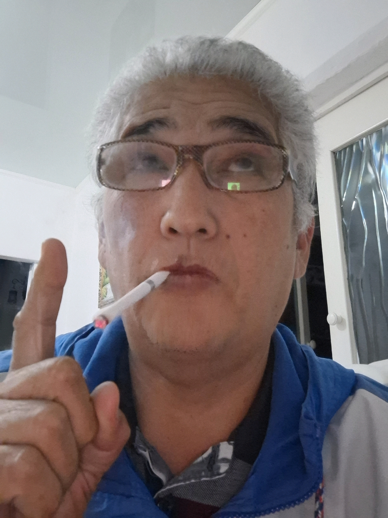 The image is a selfie of a middle-aged person with grey hair and glasses. They are holding a lit cigarette in their mouth and have their index finger raised. The person is wearing a blue jacket over a patterned shirt. The background appears to be indoors, with a white wall and a door with textured glass visible on the right side. The lighting suggests it could be daytime, but the indoor setting makes it difficult to determine the exact time of day or weather. The person's expression is neutral. There is no visible text in the image. The location context is Almaty, Kazakhstan.