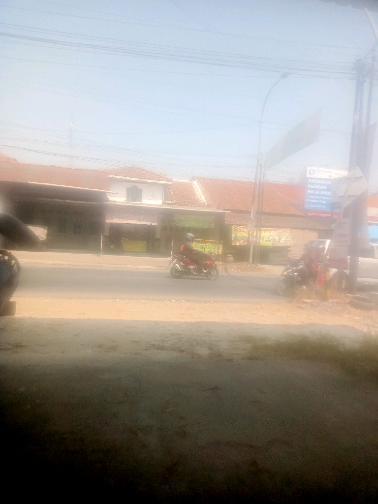 This is a photograph taken on a street in Lebaksiu, Indonesia, on a hazy day. A red motorcycle is being ridden on the asphalt road. To the left of the road are buildings with brown tile roofs, some appearing to be shops. A person is visible on the motorcycle. Another motorcycle, black in color, is parked on the right side of the road, close to a utility pole. There is a sign on a pole with blue and white text, partially obscured. The ground in the foreground is dusty and uneven. Power lines are visible stretching across the sky.