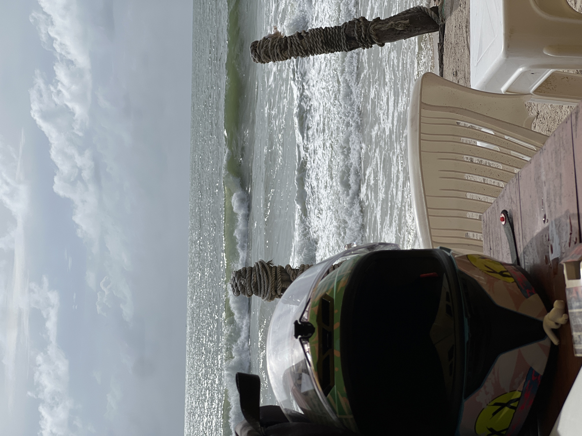 A high-angle, tilted outdoor shot features a motorcycle helmet on a table, with a beach and ocean visible in the background. The sky is predominantly gray with some scattered clouds. The ocean water is choppy with visible waves, and there are two wooden posts wrapped with rope near the shoreline. A white plastic chair sits near the table with the helmet. The helmet is black and patterned with neon pink and yellow designs. The table surface is made of dark brown wood. The image's location is in the city of Unknown, Unknown.
