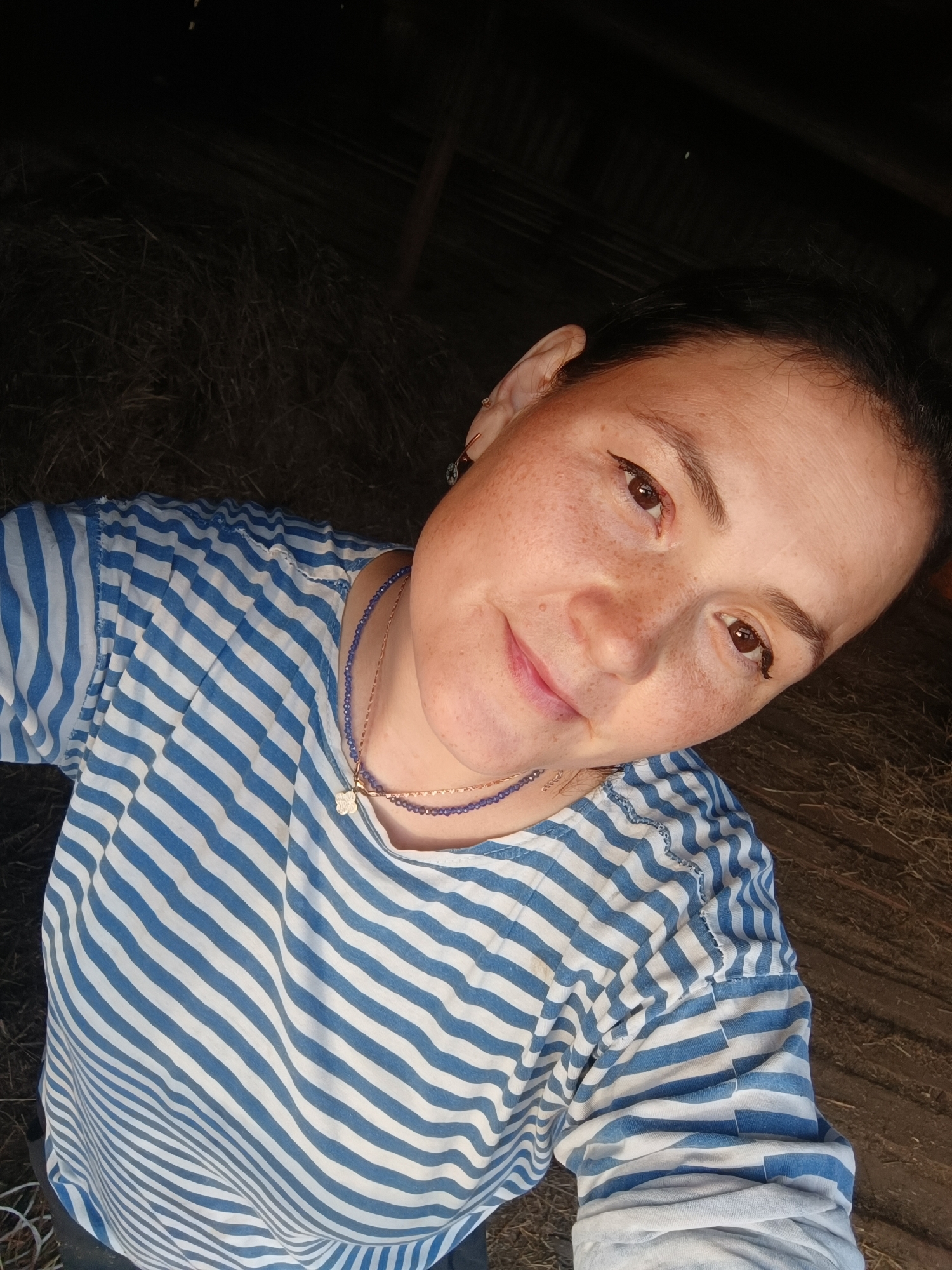 A medium shot shows a woman with freckles and brown eyes, wearing a blue and white striped t-shirt and two necklaces. She is in a dimly lit, rustic environment with straw visible in the background. The woman is angled towards the camera, with her head tilted slightly. Her hair is dark and pulled back. The lighting suggests she is indoors, possibly in a barn or storage area.