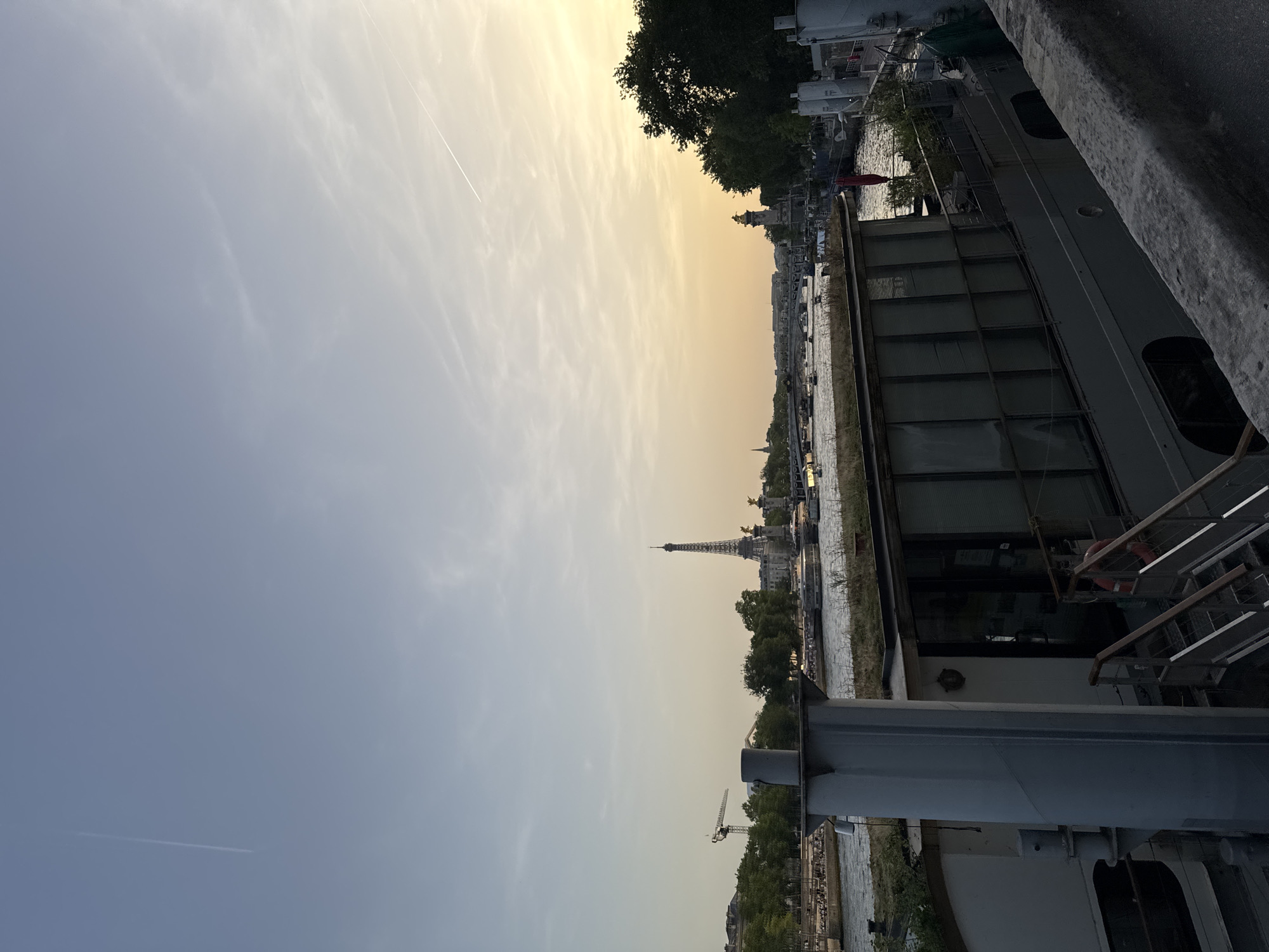 The image shows a view of the Eiffel Tower in Paris, France, from what appears to be a boat or a structure along the Seine River. The sky is a mix of pale blue and a golden hue, suggesting either sunrise or sunset. A contrail is visible in the sky. In the foreground, there is a portion of a boat or building with metallic and grey elements. Trees line the riverbank. Other buildings and structures are visible along the river.