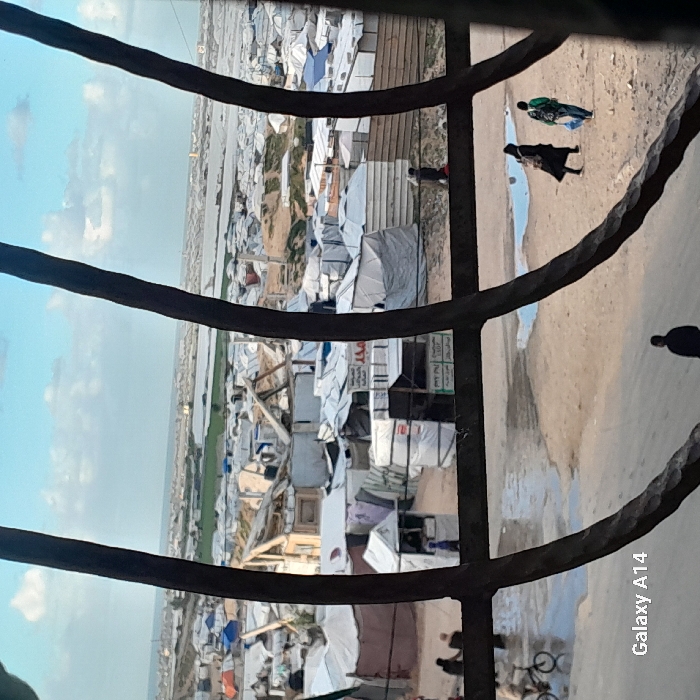 The image presents an elevated view of a sprawling displacement camp in Khan Yunis, Palestinian Territory, observed through four curved, dark, textured metal bars of a window or grille.
The immediate foreground is dominated by the dark metal bars. At the bottom left, a "Galaxy A14" watermark is present.
Below the main horizontal bar, the ground is unpaved, sandy earth with multiple puddles of standing water reflecting the sky. On the left, a person is seen riding a bicycle through a puddle, while two other figures stand nearby. On the right, two individuals walk away from the viewer; one wears dark clothing and a head covering, and the other carries a light-colored bag. A lone figure in dark clothing is partially visible at the bottom center, facing away.
In the midground, beyond the unpaved area, a dense collection of makeshift structures, tents, and shelters extends into the distance. Many are constructed from white or light-colored tarpaulins. Several more substantial, temporary buildings with exposed wooden framing and corrugated metal are visible, some appearing partially damaged or deconstructed. Arabic text is discernible on signs attached to some structures, including one that reads "محمد" (Mohammed). Makeshift fences or barriers divide sections of the camp.
The background reveals further layers of the camp stretching towards the horizon. Beyond the camp, a strip of green agricultural land, possibly containing greenhouses, is visible. The Mediterranean Sea forms the distant horizon under a sky with scattered white clouds. A tall, slender structure, potentially a minaret or tower, is faintly visible on the distant horizon line, slightly right of center.