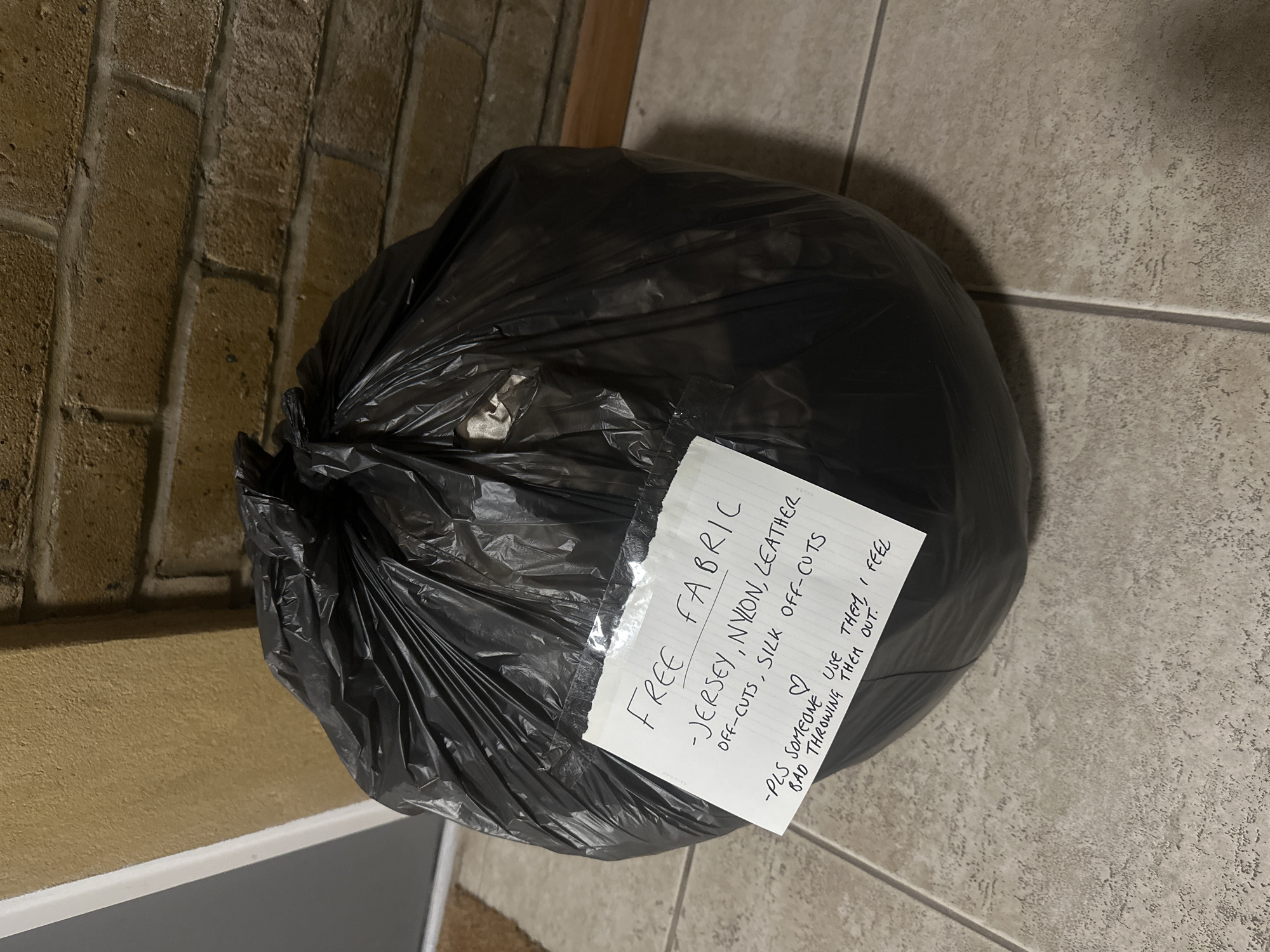 A black garbage bag filled with fabric scraps is sitting on a tiled floor near a brick wall. A note taped to the bag reads "FREE FABRIC - JERSEY, NYLON, LEATHER OFF-CUTS, SILK OFF-CUTS -PLS SOMEONE ♡ USE THEM, I FEEL BAD THROWING THEM OUT." The specific location in London, United Kingdom is not discernible from the image.
