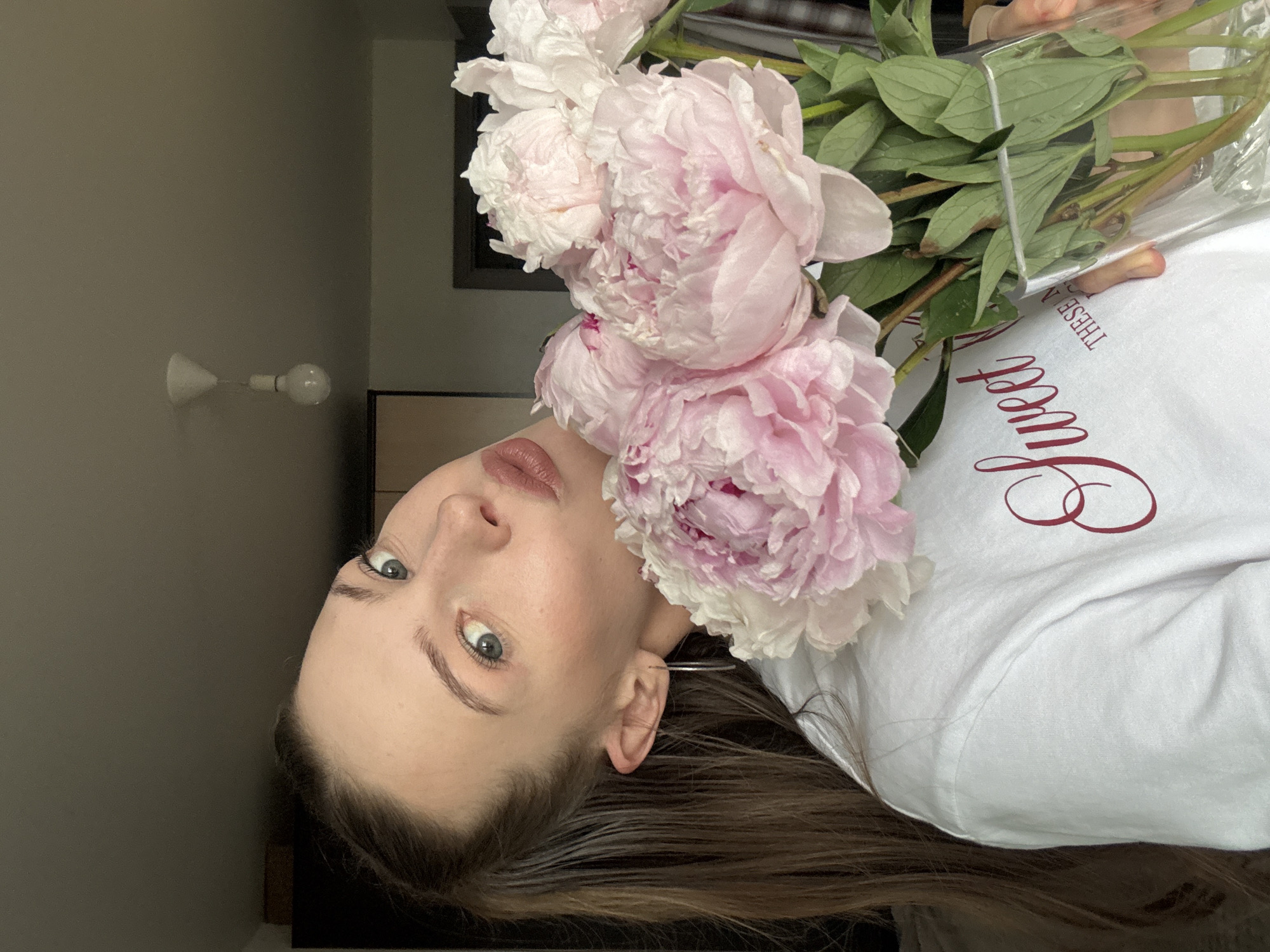 The media file shows a close-up, upside-down shot of a young woman holding a bouquet of pink peonies. The woman has fair skin, blue-green eyes, and long, dark brown hair. She is wearing a white t-shirt with red script text that says "Sweet." The background is a muted gray wall and a dark interior. A wall-mounted light fixture is visible on the left side of the image. The lighting suggests it is daytime indoors. The overall mood appears serene and intimate. The text on the shirt is a notable detail. The location context provided is Perm, Russia.