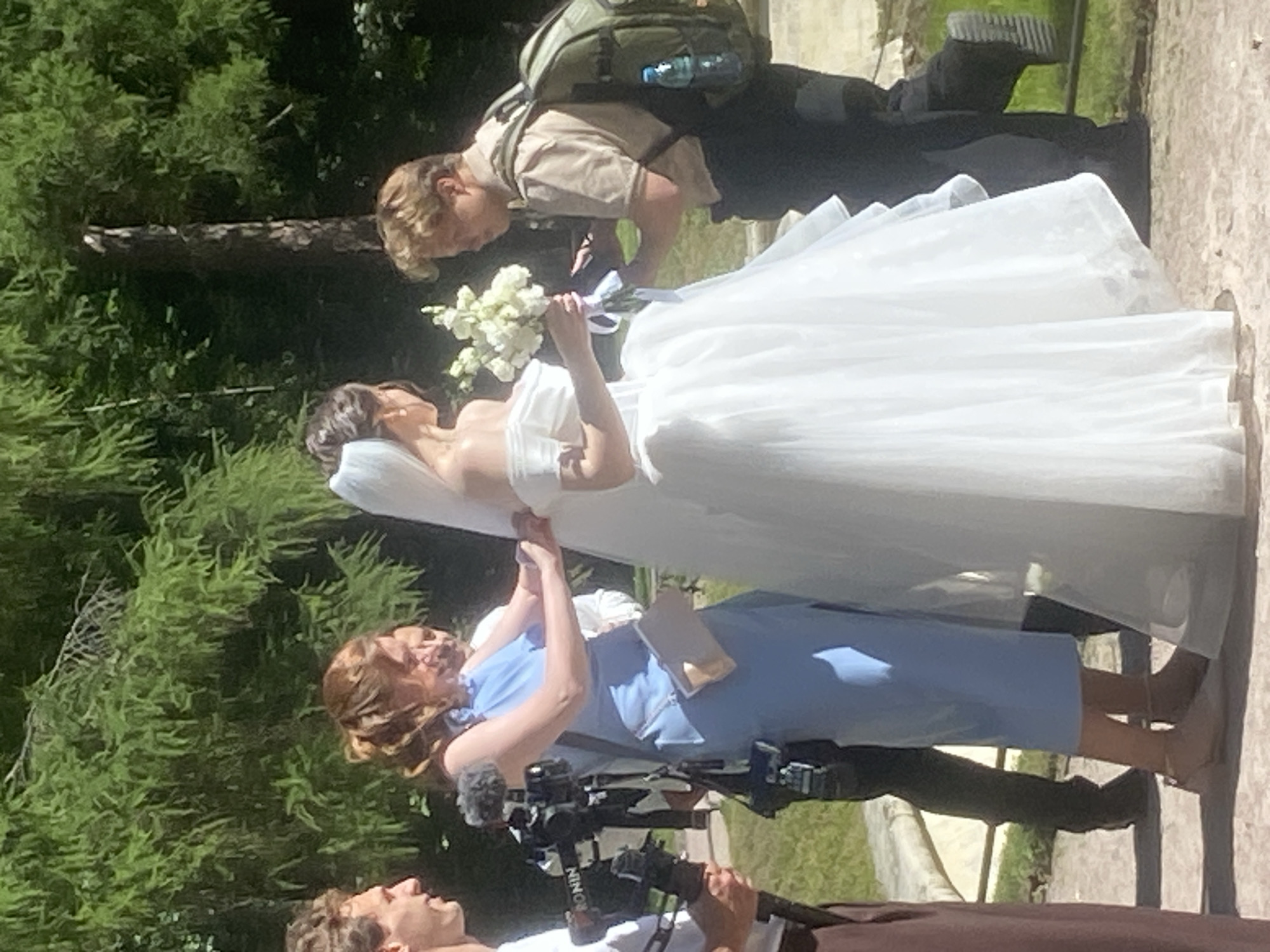 This image captures a wedding scene outdoors, possibly in the city of Pushkin, Russia, given the architectural hints and lush greenery suggestive of a park or estate. In the center, a bride in a flowing white off-the-shoulder wedding dress with a veil stands holding a bouquet of white flowers. To her left, a woman in a light blue dress is adjusting the bride's attire. To the left of the woman in blue, a person holding a professional camera on a gimbal is visible. To the right of the bride, a man wearing a tan shirt and dark pants, with a backpack, appears to be interacting with the bride. The background is dominated by dense green foliage of trees, suggesting a sunny day with dappled light. A stone bench is partially visible in the lower left of the frame.