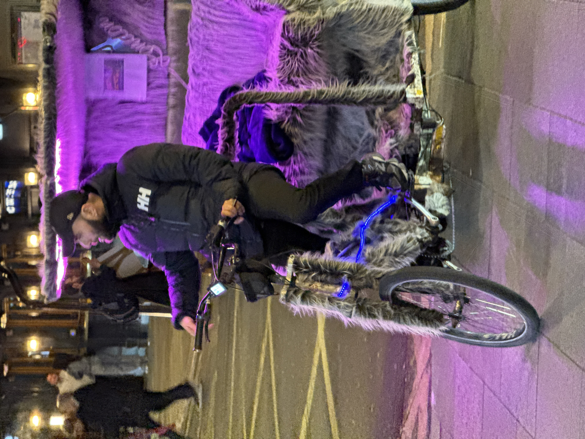 A man rides a modified bicycle on a city street. The bicycle is covered in faux fur, with blue LED lights attached. The man is wearing a black jacket with white lettering and a black cap. The street is marked with yellow lines. A group of people is visible in the background. The location is identified as London, United Kingdom.