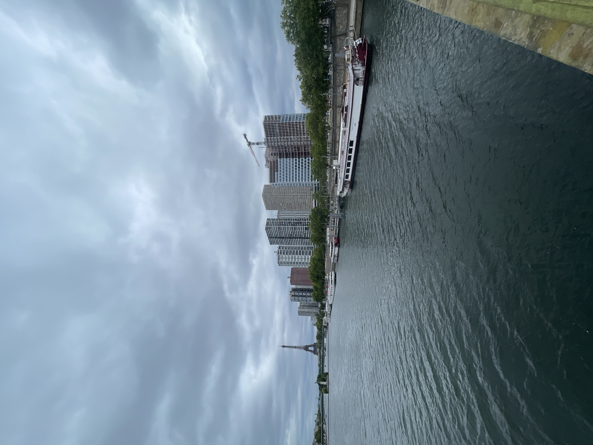 The image captures a view of the River Seine in Paris, France, under a cloudy sky. In the distance, the Eiffel Tower stands tall on the left side of the frame. Along the right bank of the river, a line of modern high-rise buildings is visible, some of which appear to be under construction, indicated by a crane. Several houseboats are moored along the riverbank. The water of the Seine is dark and rippled, suggesting a calm but overcast day. The perspective is from a bridge or elevated viewpoint looking across the river towards the city skyline.